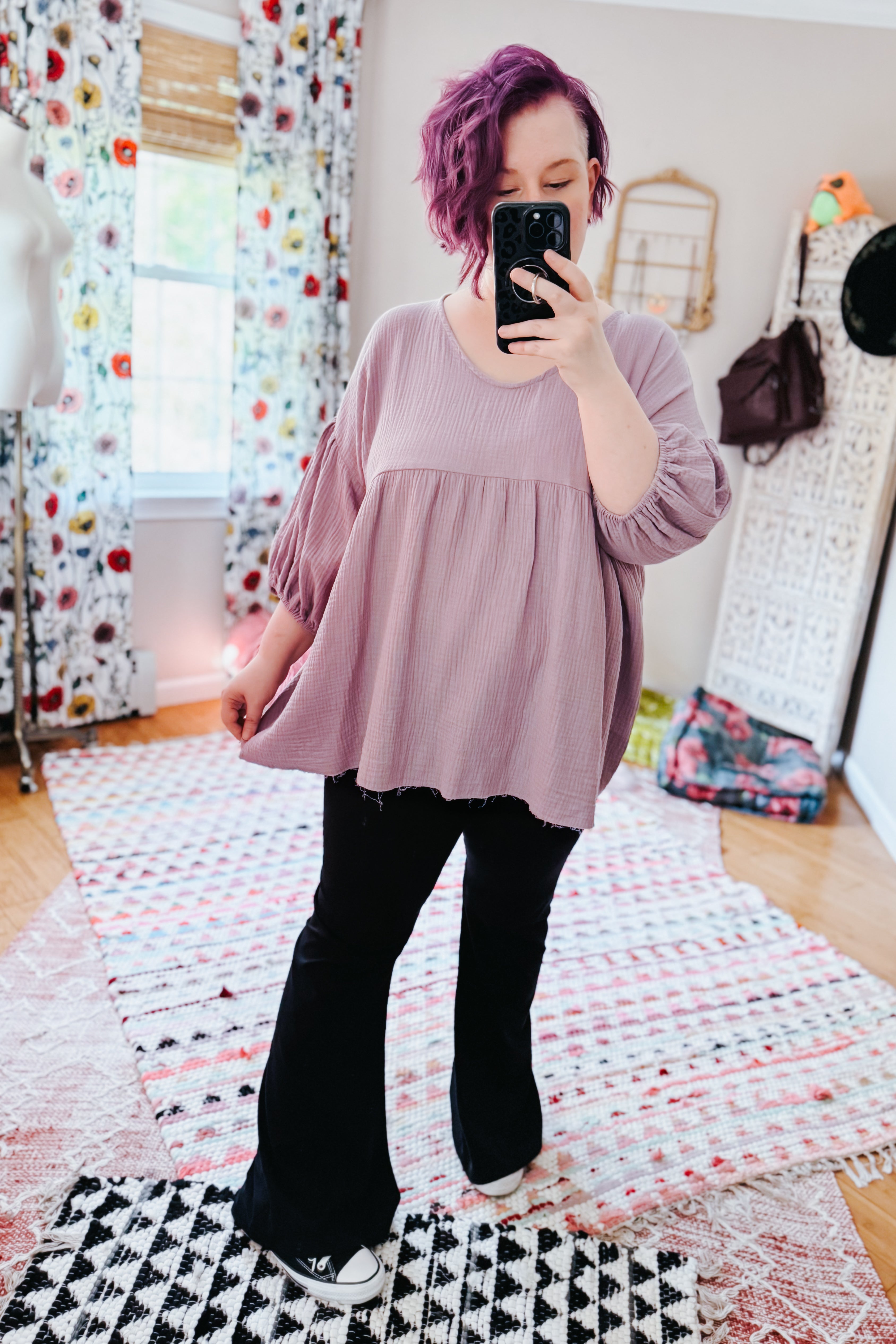 Woman with purple hair wearing a purple bubble sleeve peasant blouse and black flared pants in a cozy eclectic room