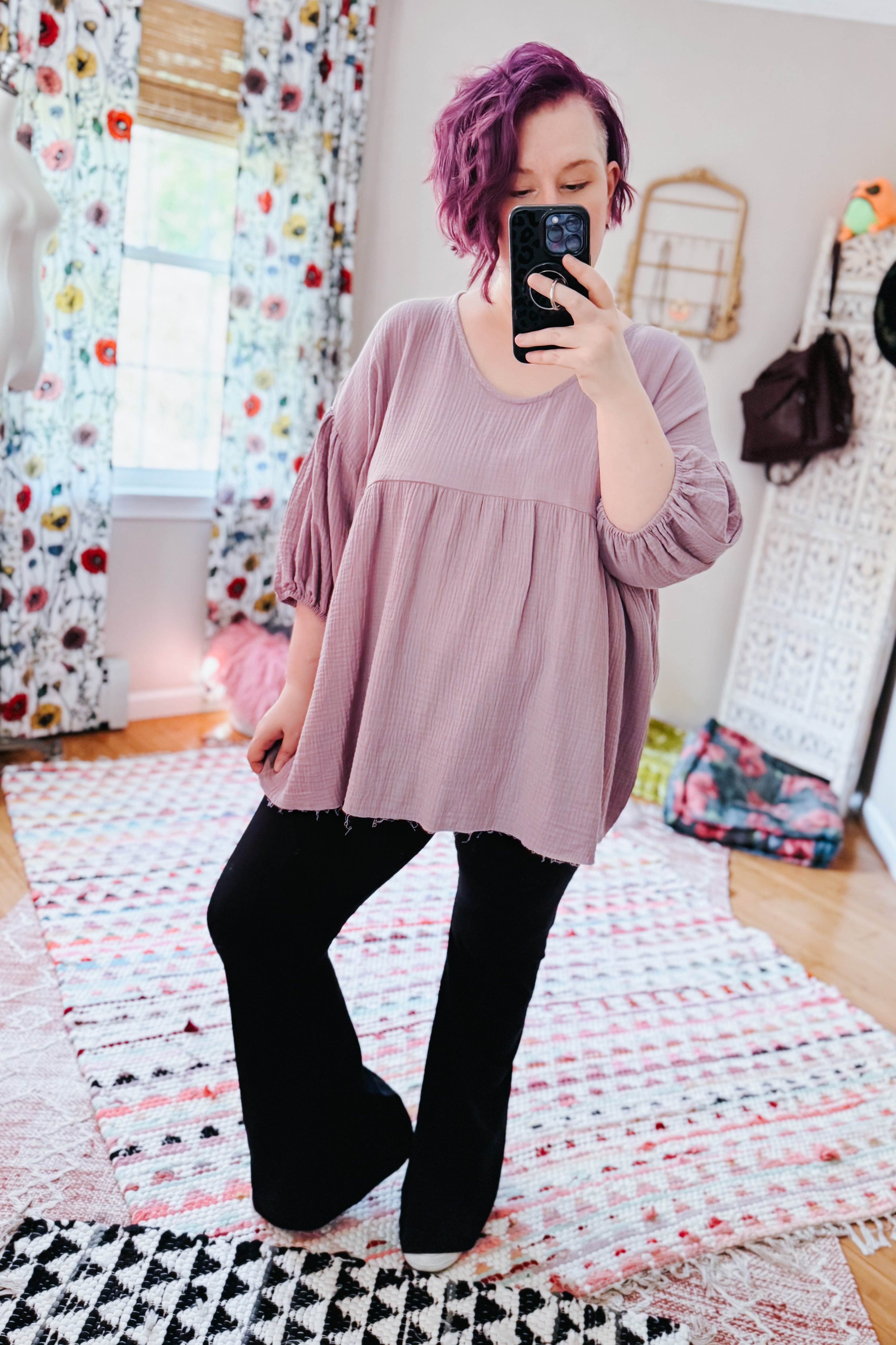 Woman with purple hair wearing a soft purple bubble sleeve blouse and black pants in a bright, eclectic, free-spirited room