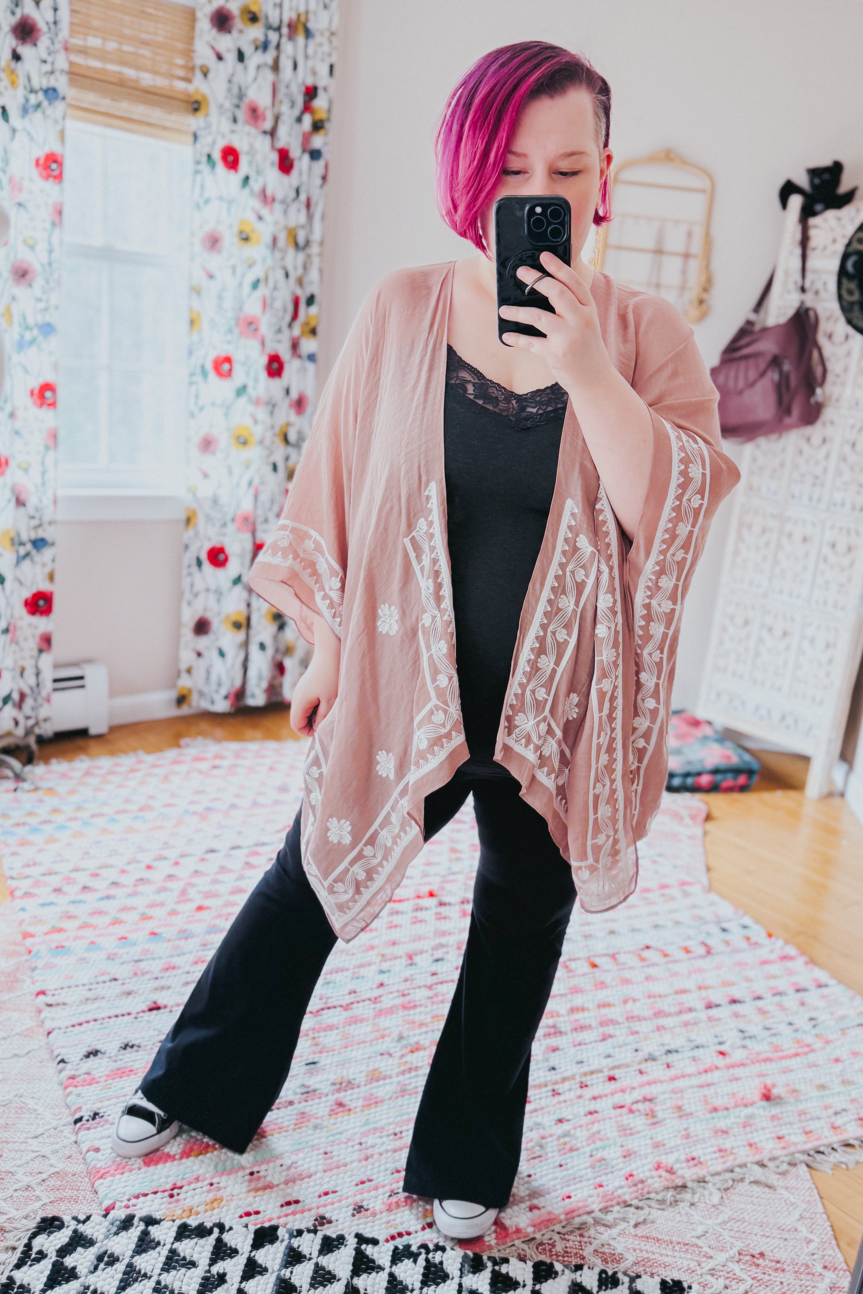 Woman wearing blush taupe layering piece with white poppy embroidery over black lace-trimmed top and wide-leg pants indoors