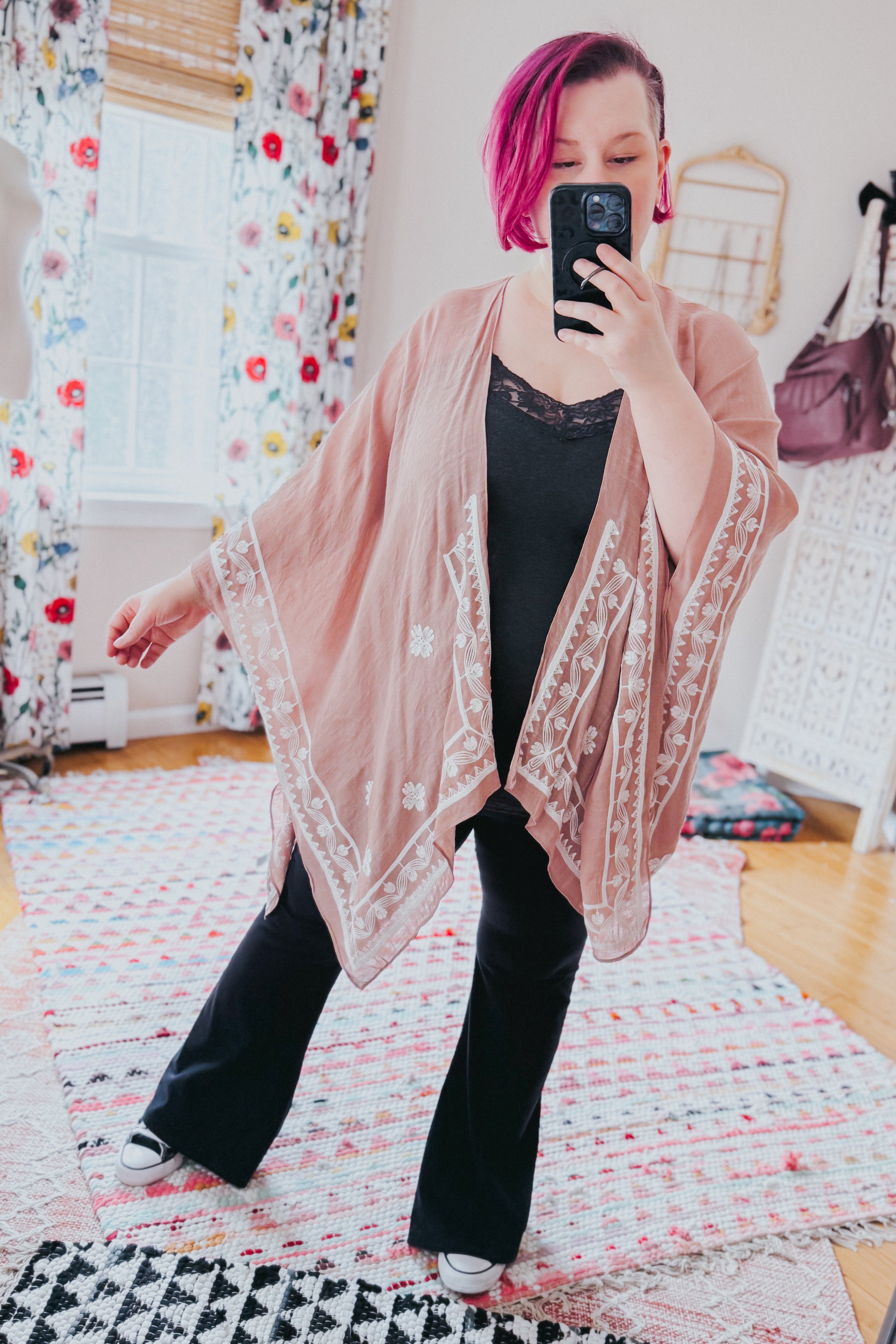 Woman with pink hair wearing blush taupe draped piece with white embroidery over black top and wide-leg pants indoors
