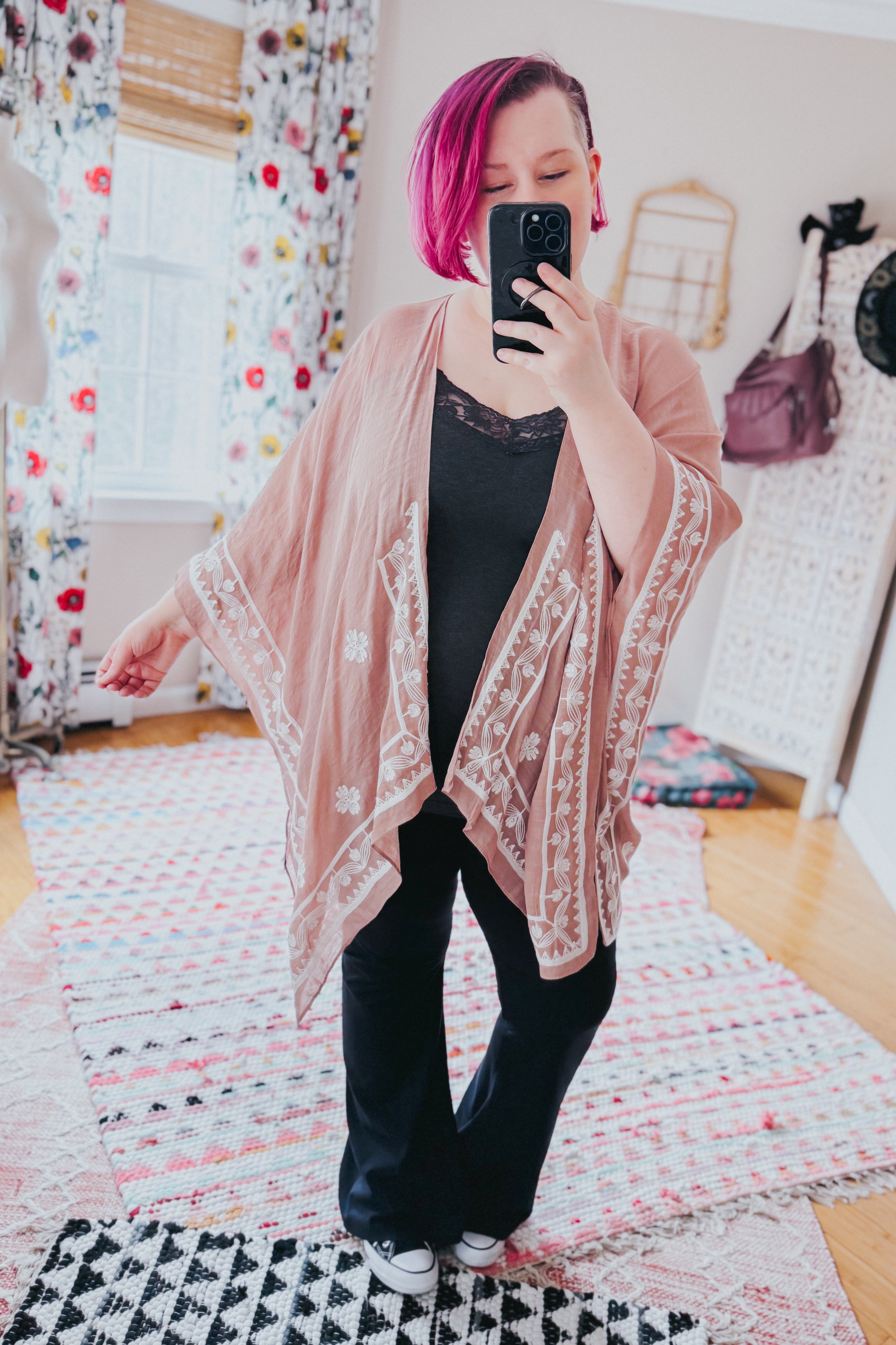 Woman with vibrant pink hair wearing blush taupe embroidered layering piece over black lace-trimmed top and pants indoors