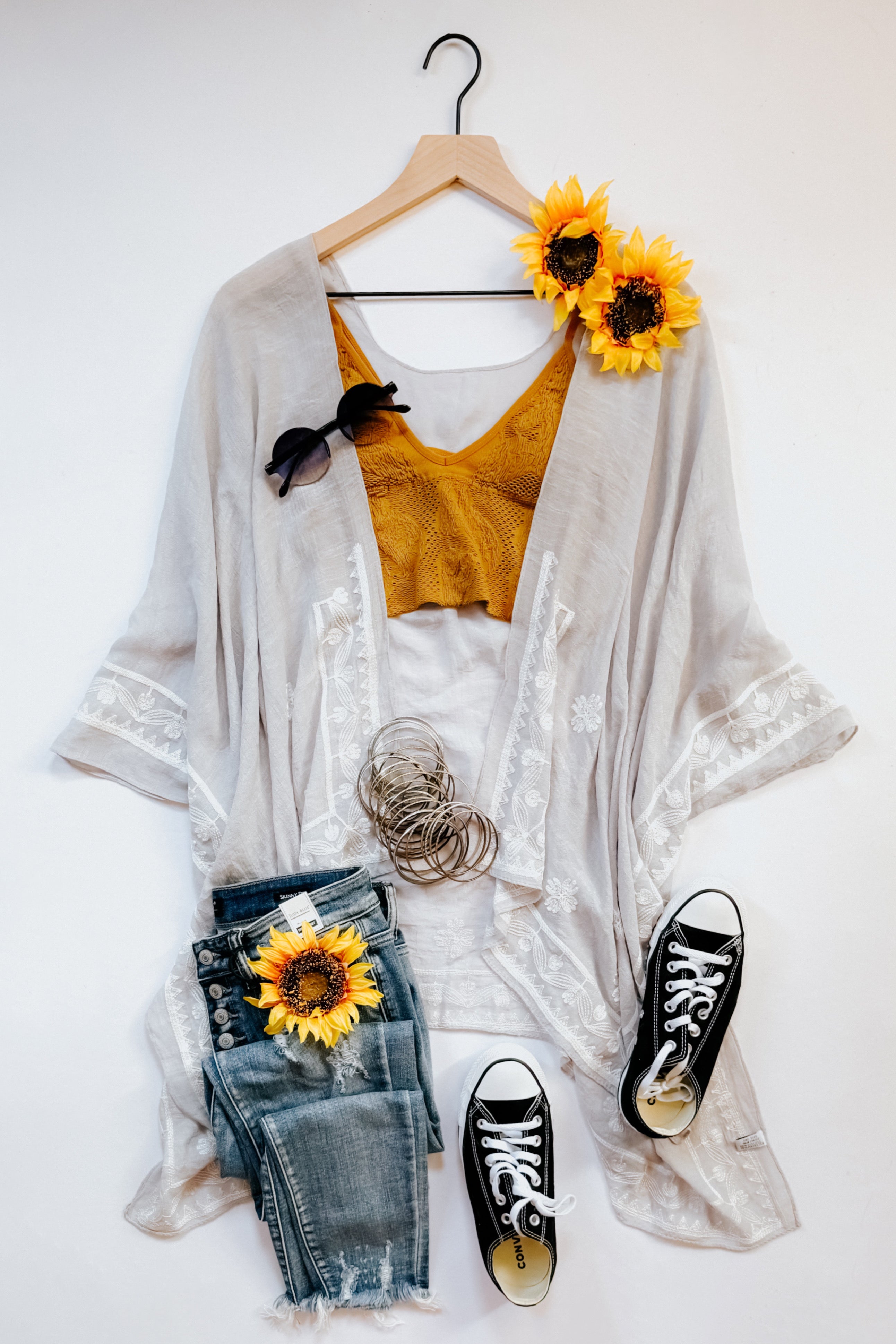 Light stone embroidered layering piece paired with mustard top, distressed denim, bracelets, black sneakers, and sunflowers on hanger.