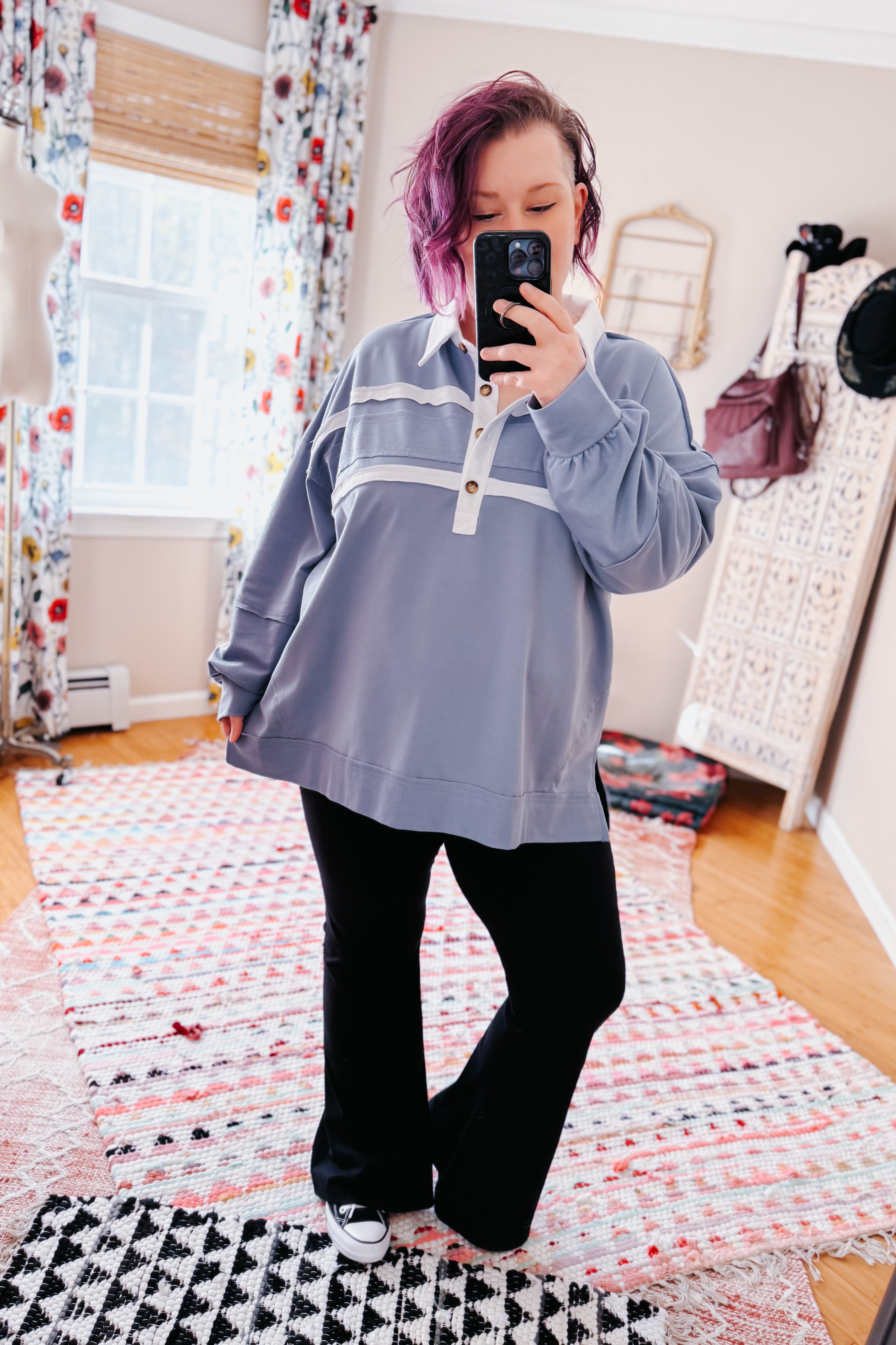Woman with purple hair wearing oversized steel blue polo top with white stripes and wooden buttons paired with black pants and sneakers taking selfie