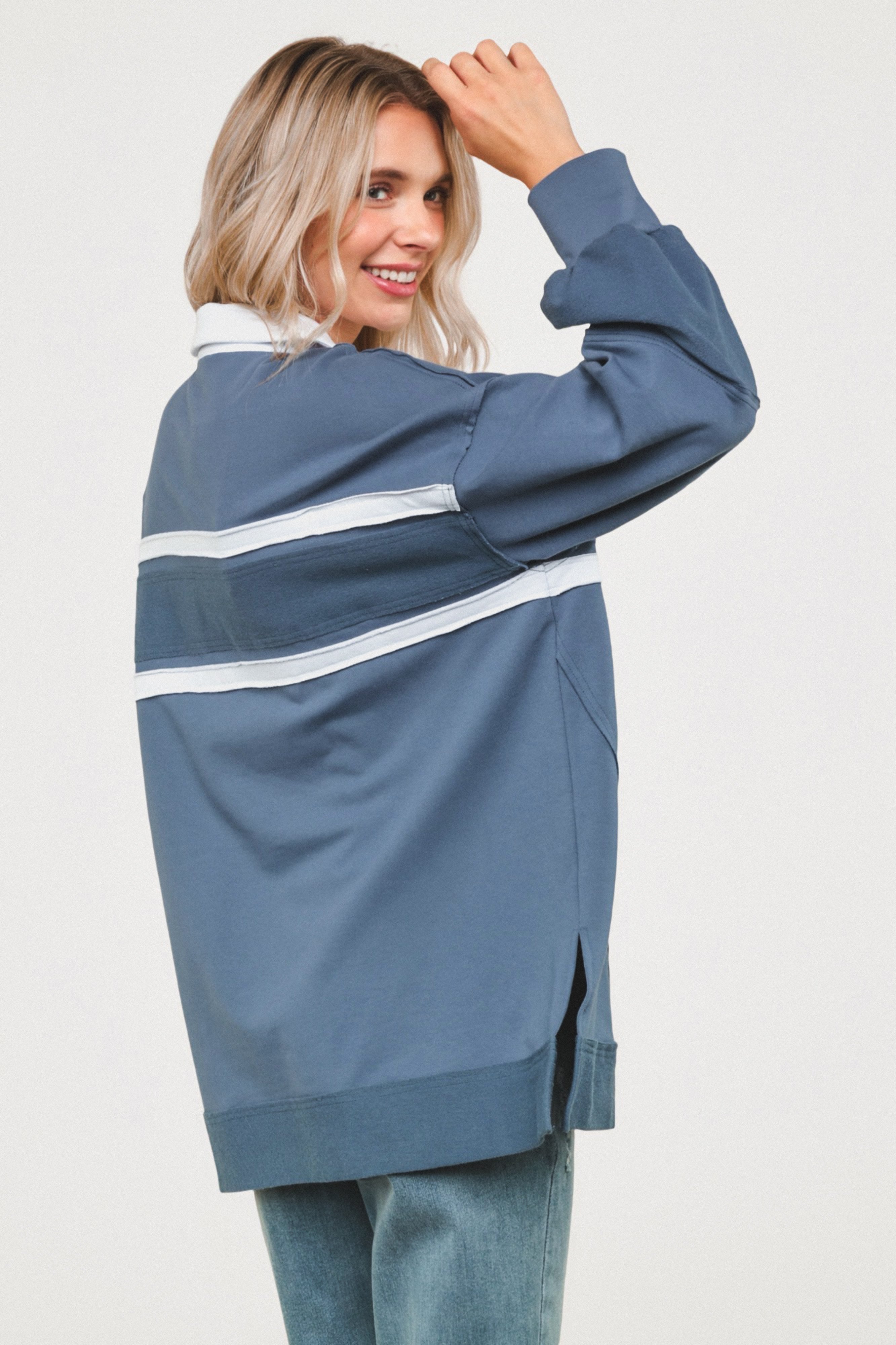 Smiling woman wearing oversized steel blue polo top with white stripes and cuffed sleeves against white backdrop