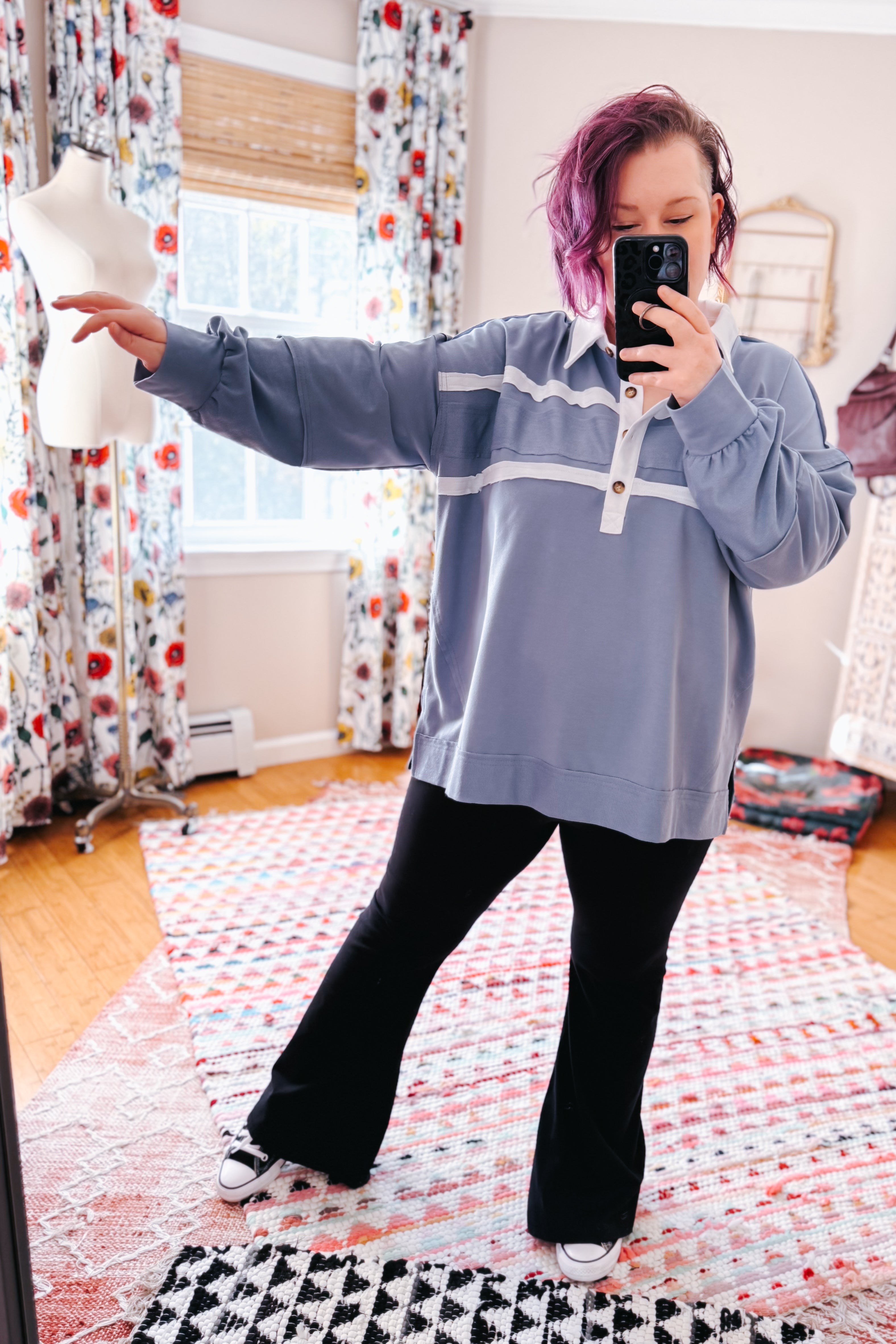 Woman with purple hair wearing oversized slate blue polo with white stripes and wooden buttons paired with black pants taking selfie