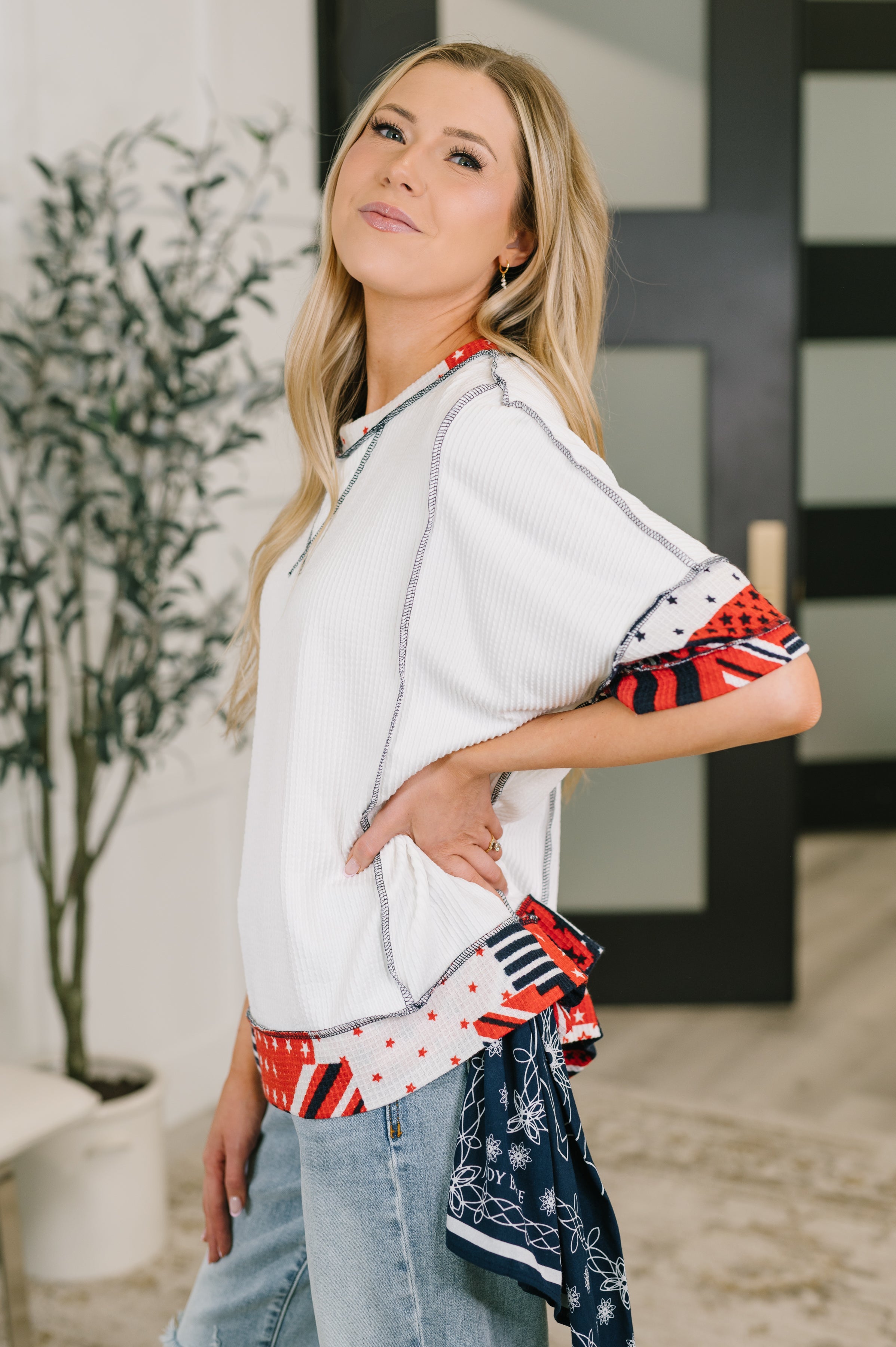 Woman wearing a cream patchwork detail top with red and navy accents paired with light denim in an eclectic indoor space
