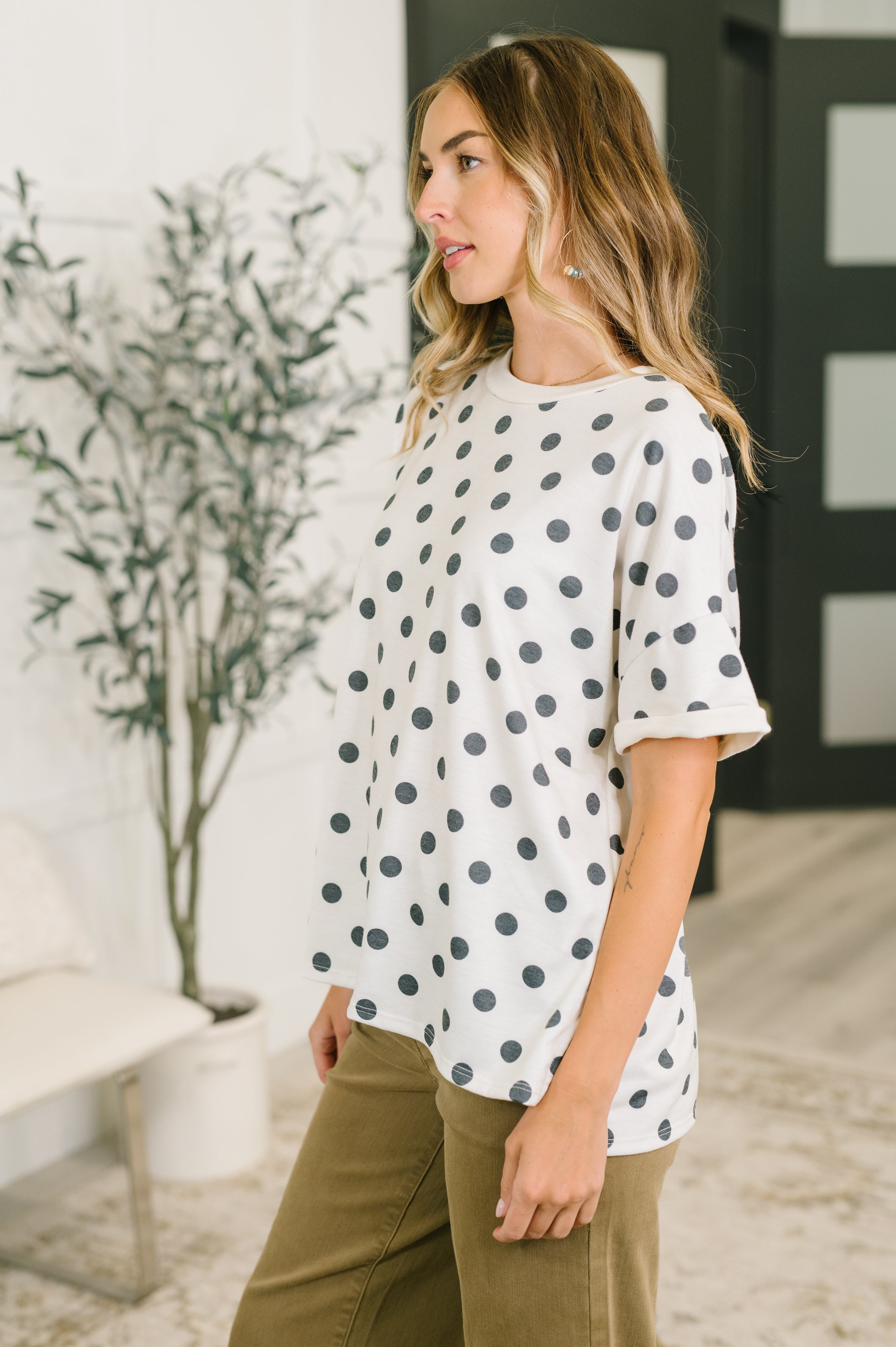Side profile of woman in ivory oversized polka dot top with rolled sleeves paired with olive pants indoors