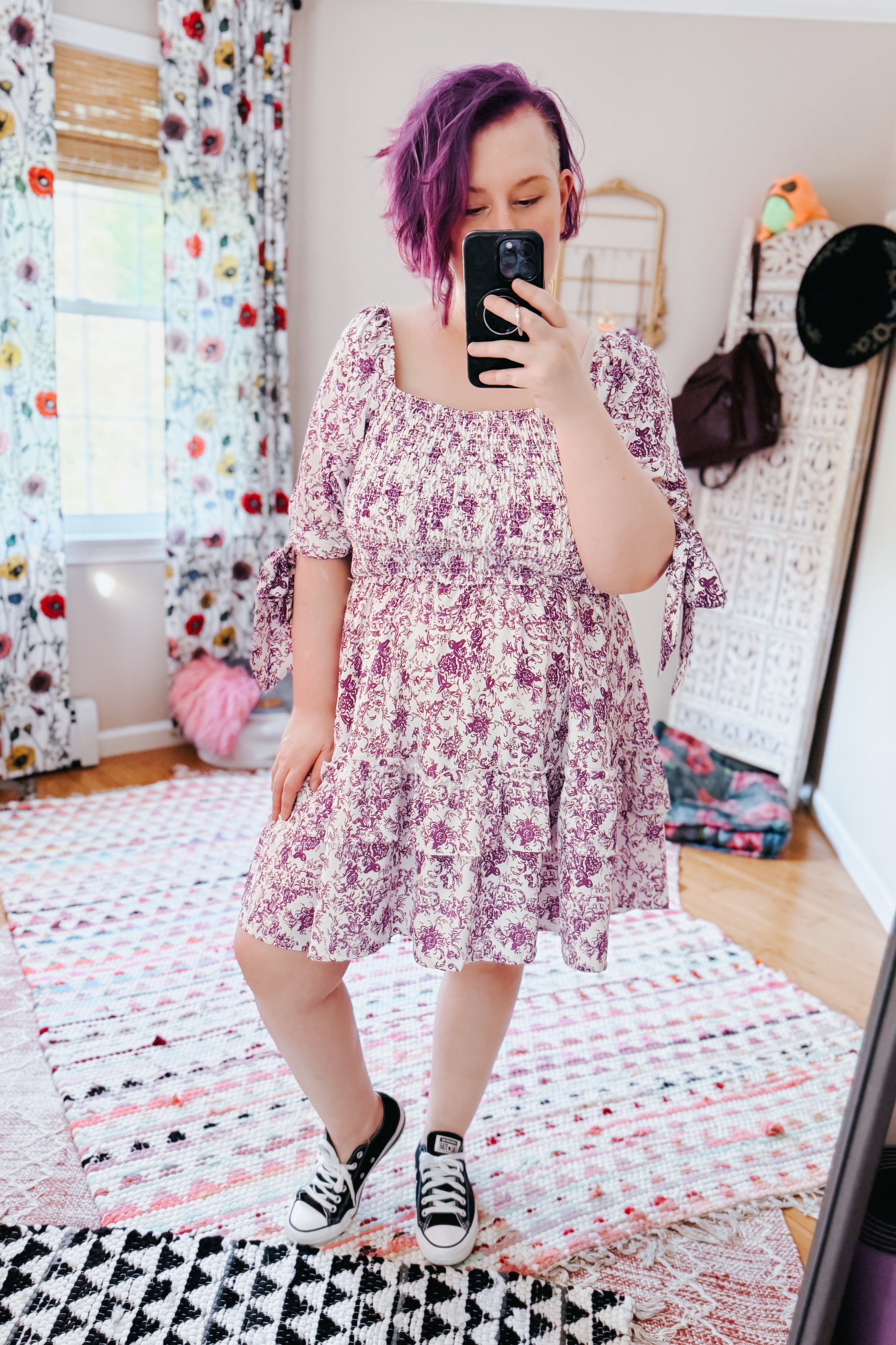 Woman with purple hair wearing a purple floral babydoll dress with puff sleeves and black sneakers in an eclectic room
