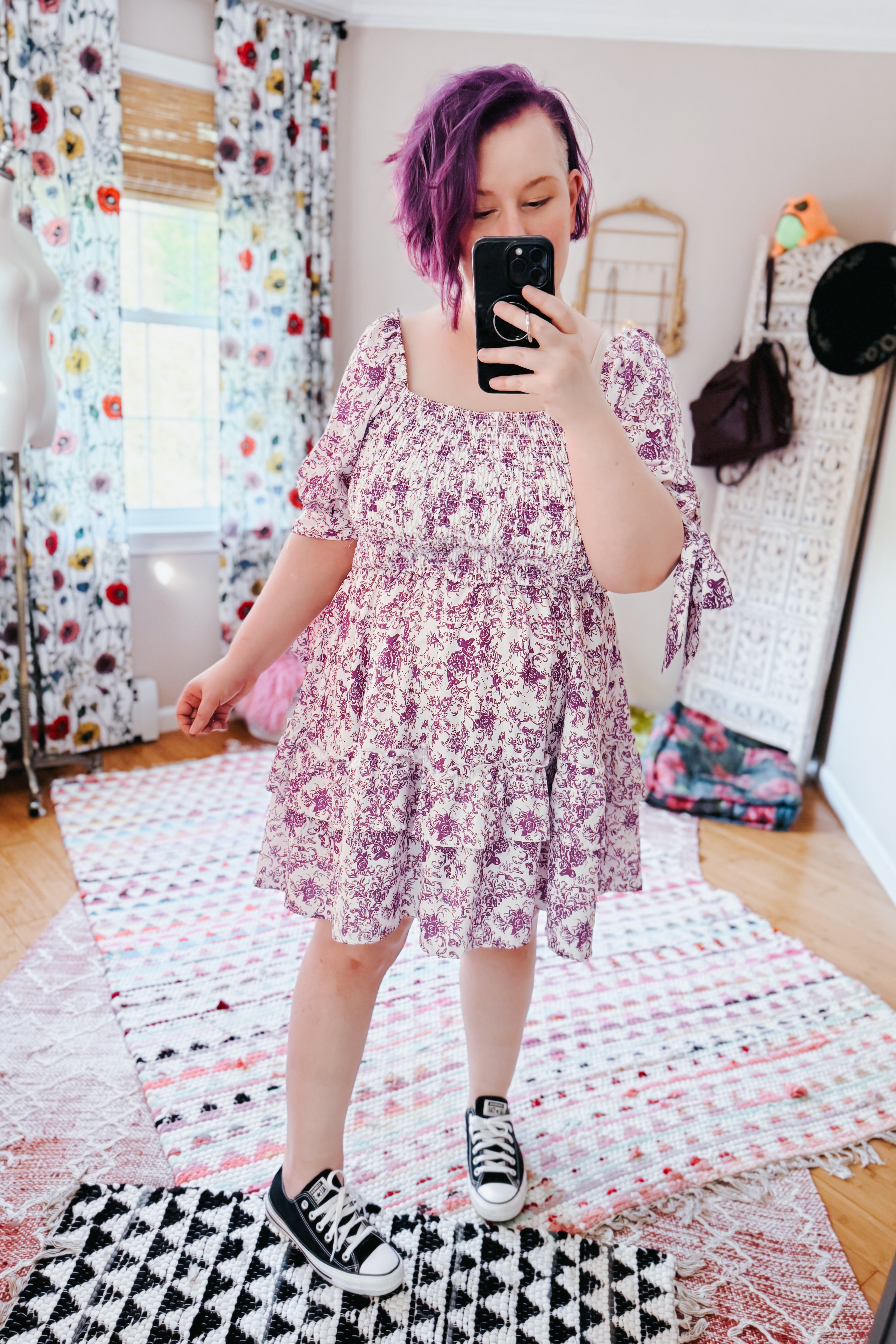 Woman with purple hair wearing a purple floral babydoll dress with puff sleeves and black sneakers in an eclectic room