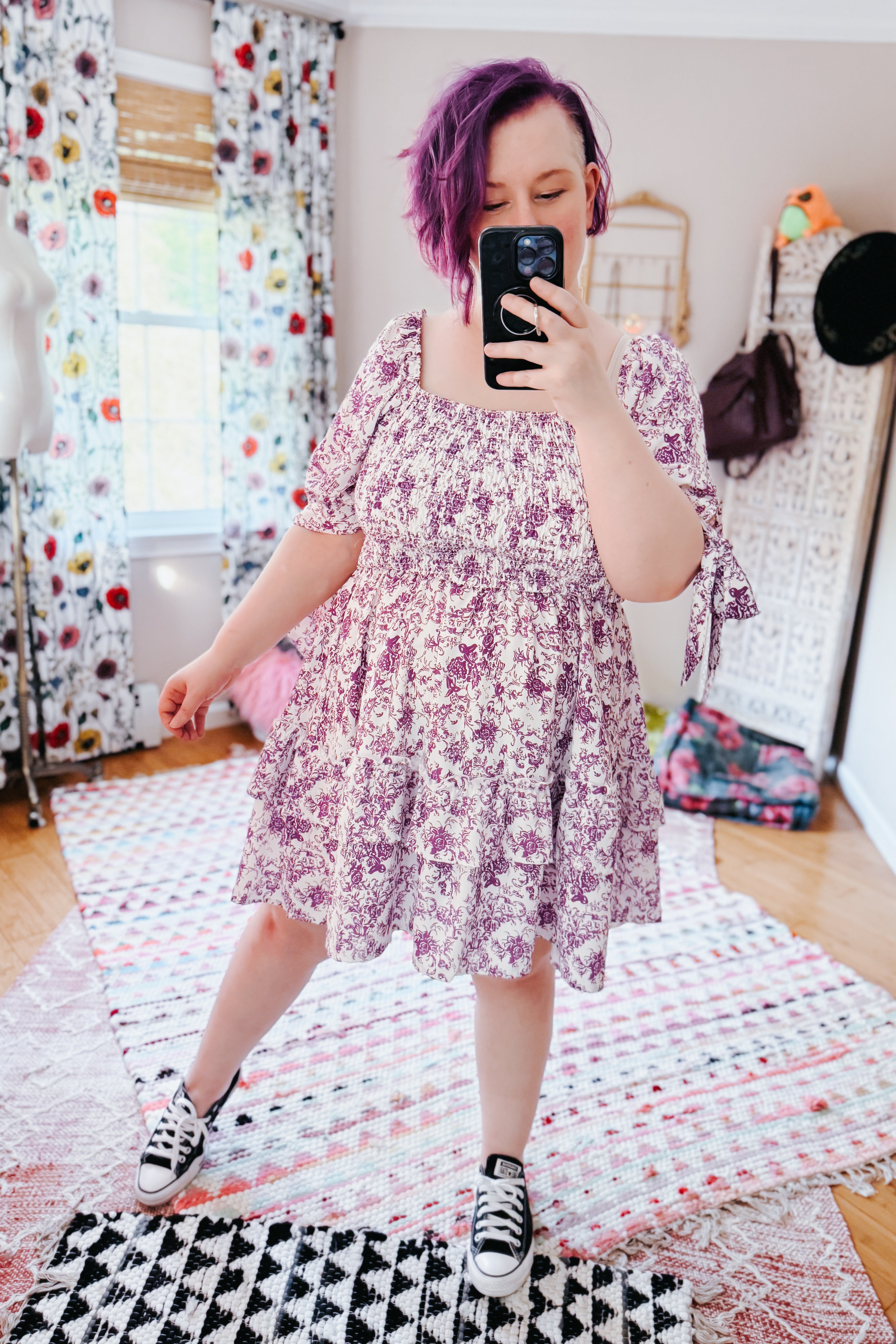 Woman with short purple hair wearing a purple floral babydoll dress with puff sleeves in an eclectic bright room