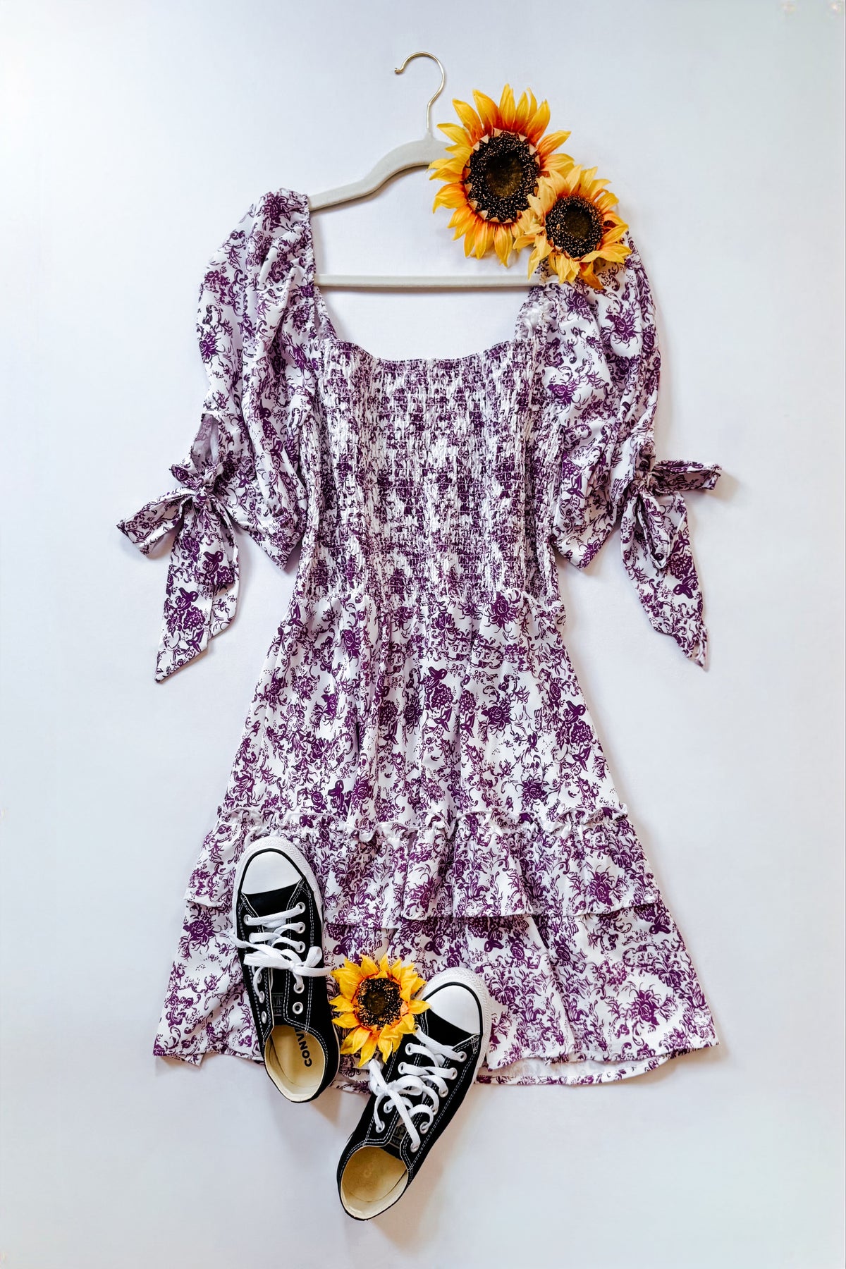 Purple floral babydoll dress with smocked bodice elastic neckline tied sleeves and ruffled tiered skirt styled with sunflowers and black sneakers
