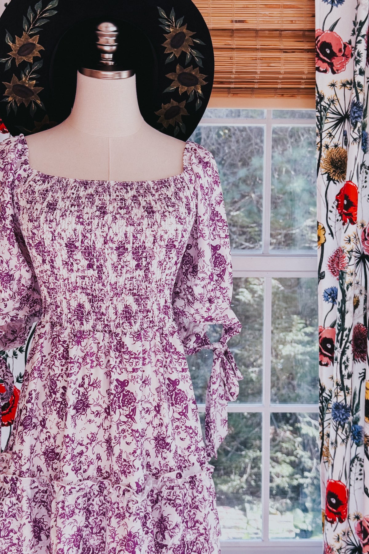 Purple floral babydoll dress with a square neckline tiered skirt and tie detail sleeves displayed on a mannequin by a window