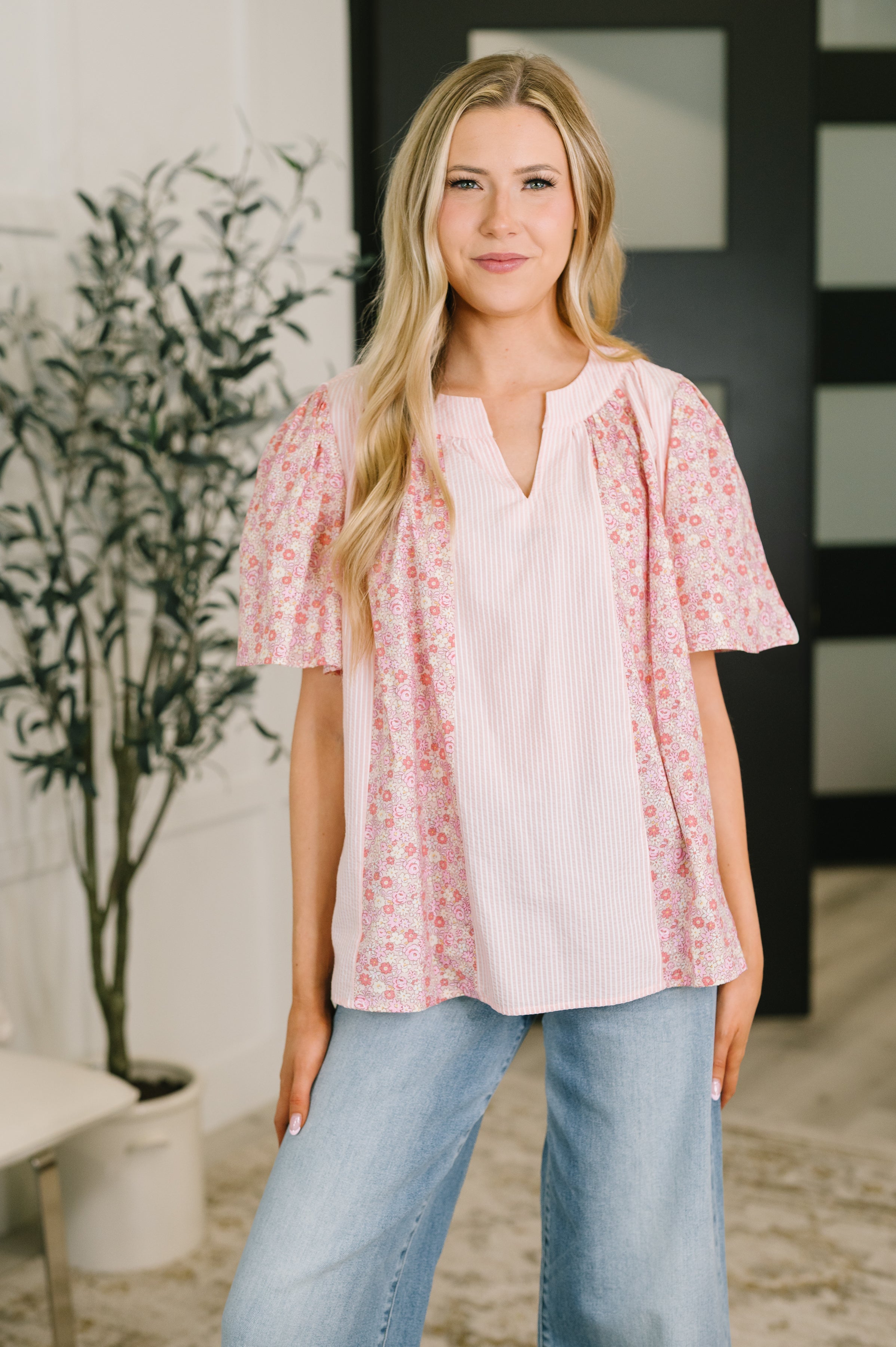 Woman wearing a pink bell sleeve blouse with striped and floral color block panels paired with light denim jeans indoors