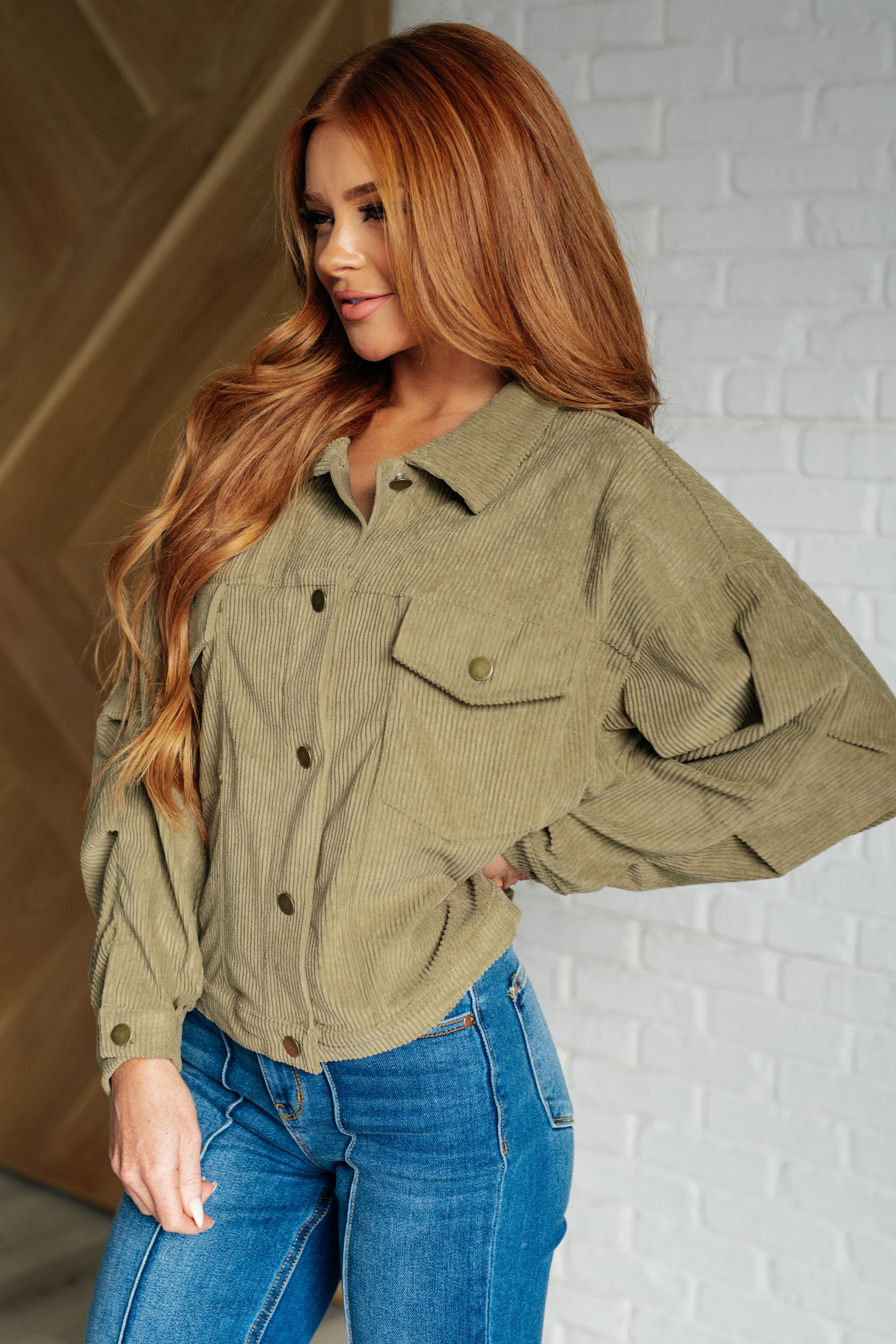 Woman with long hair wearing an olive corduroy jacket with snap buttons and blue jeans indoors, smiling softly