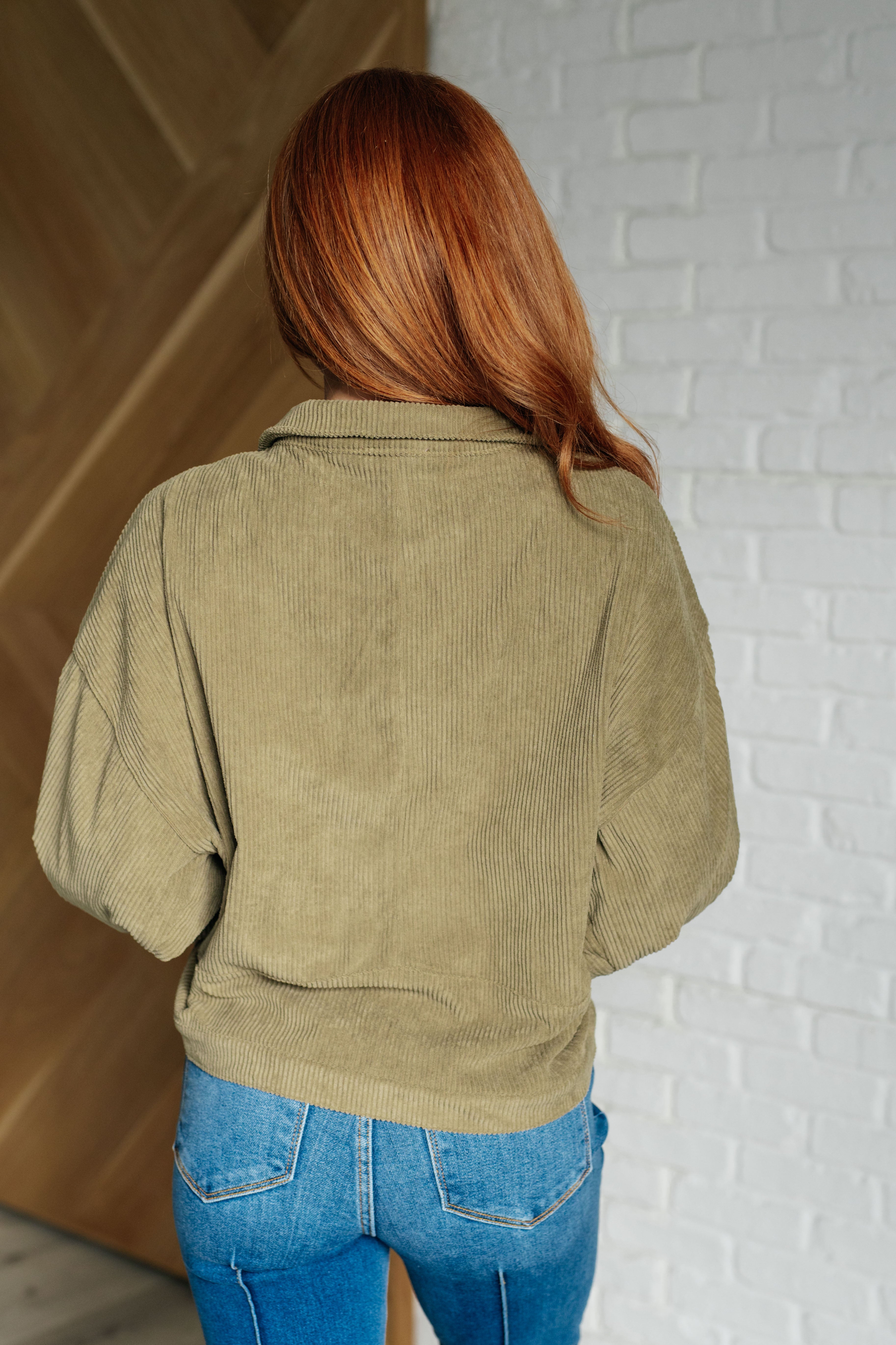 Back view of woman with red hair wearing an olive corduroy jacket and blue jeans indoors
