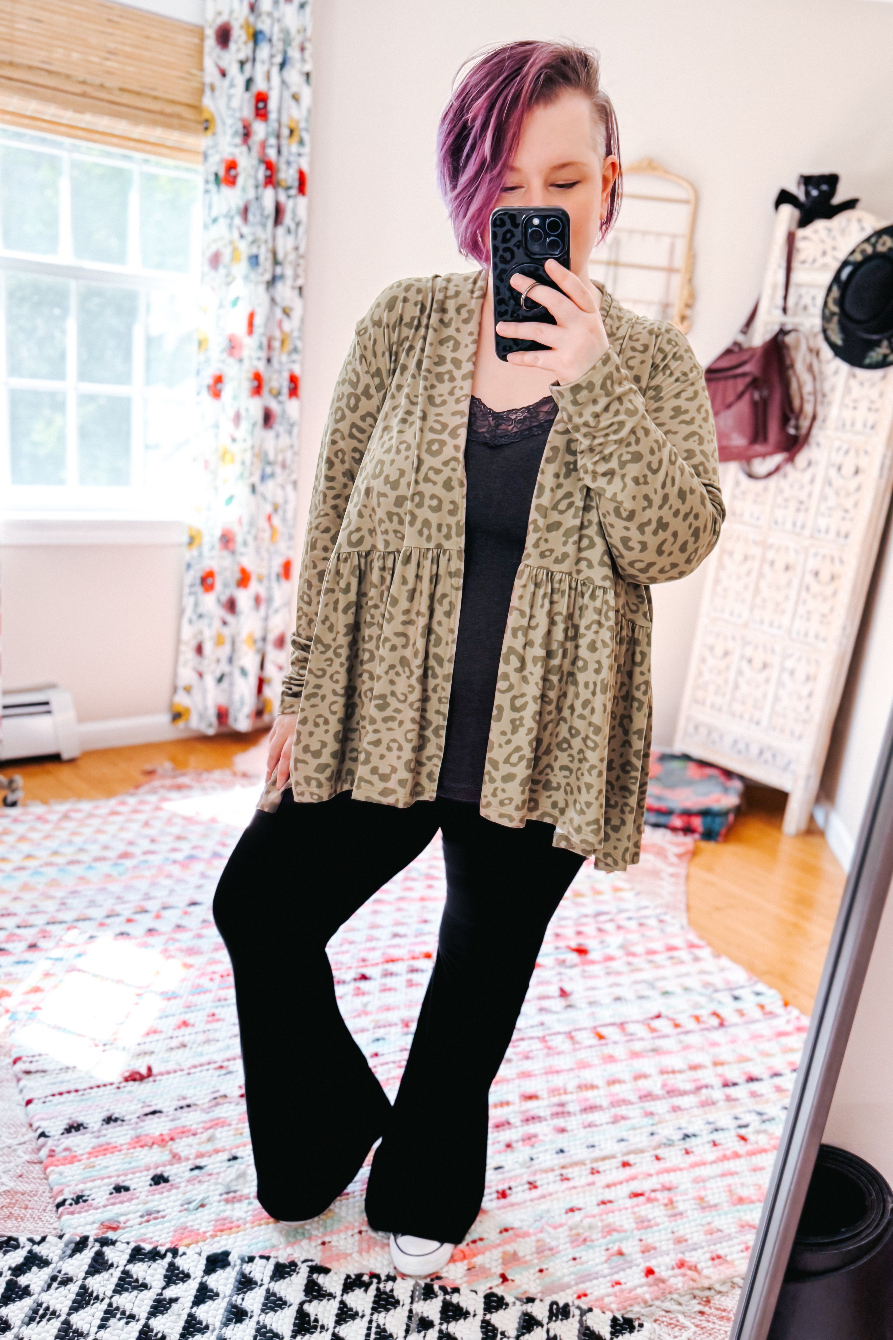 Woman with purple hair wearing green leopard print peplum open cardigan over black top and pants in bright eclectic room