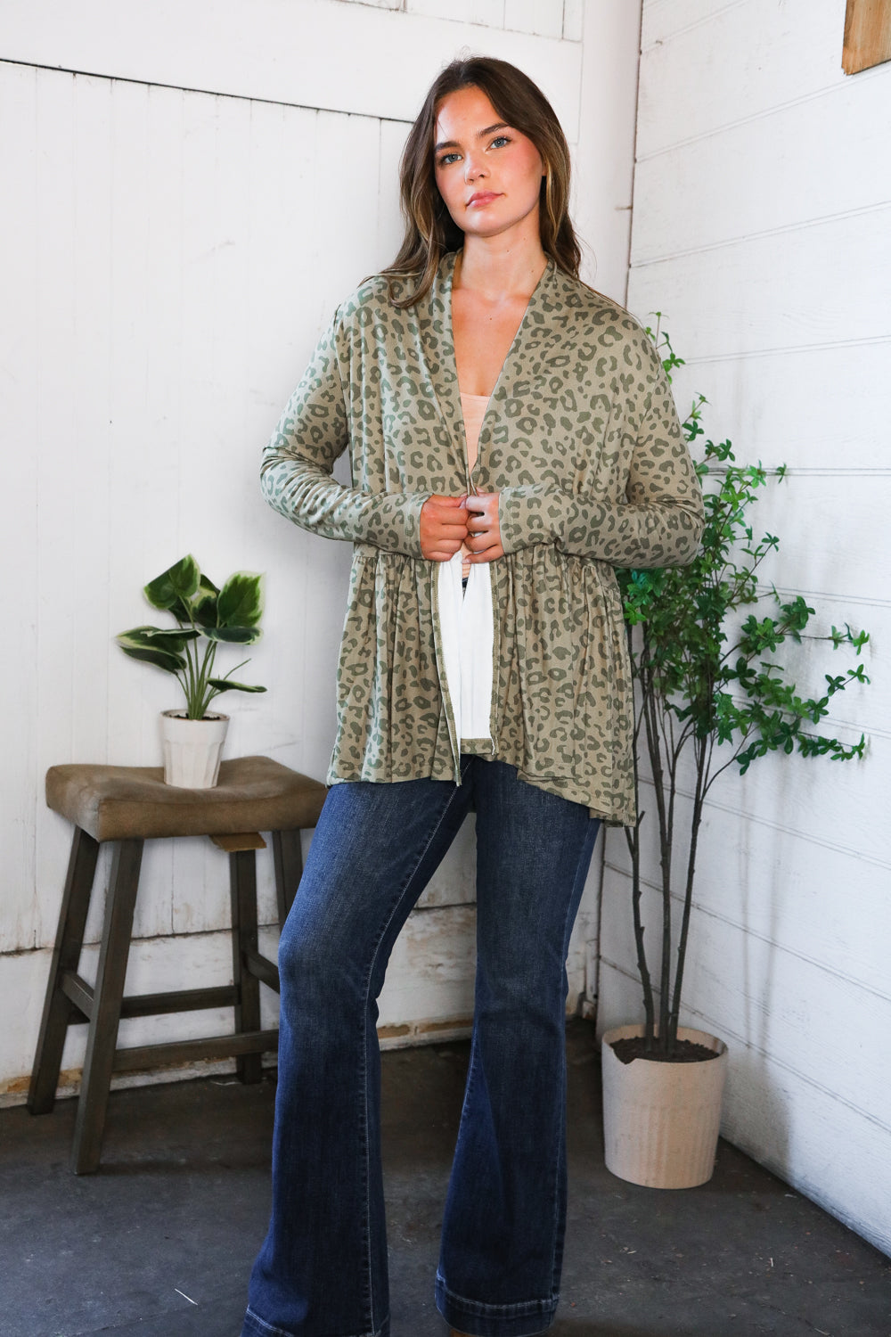 Woman wearing green leopard print peplum cardigan layered over light top in eclectic space with plants and stool nearby