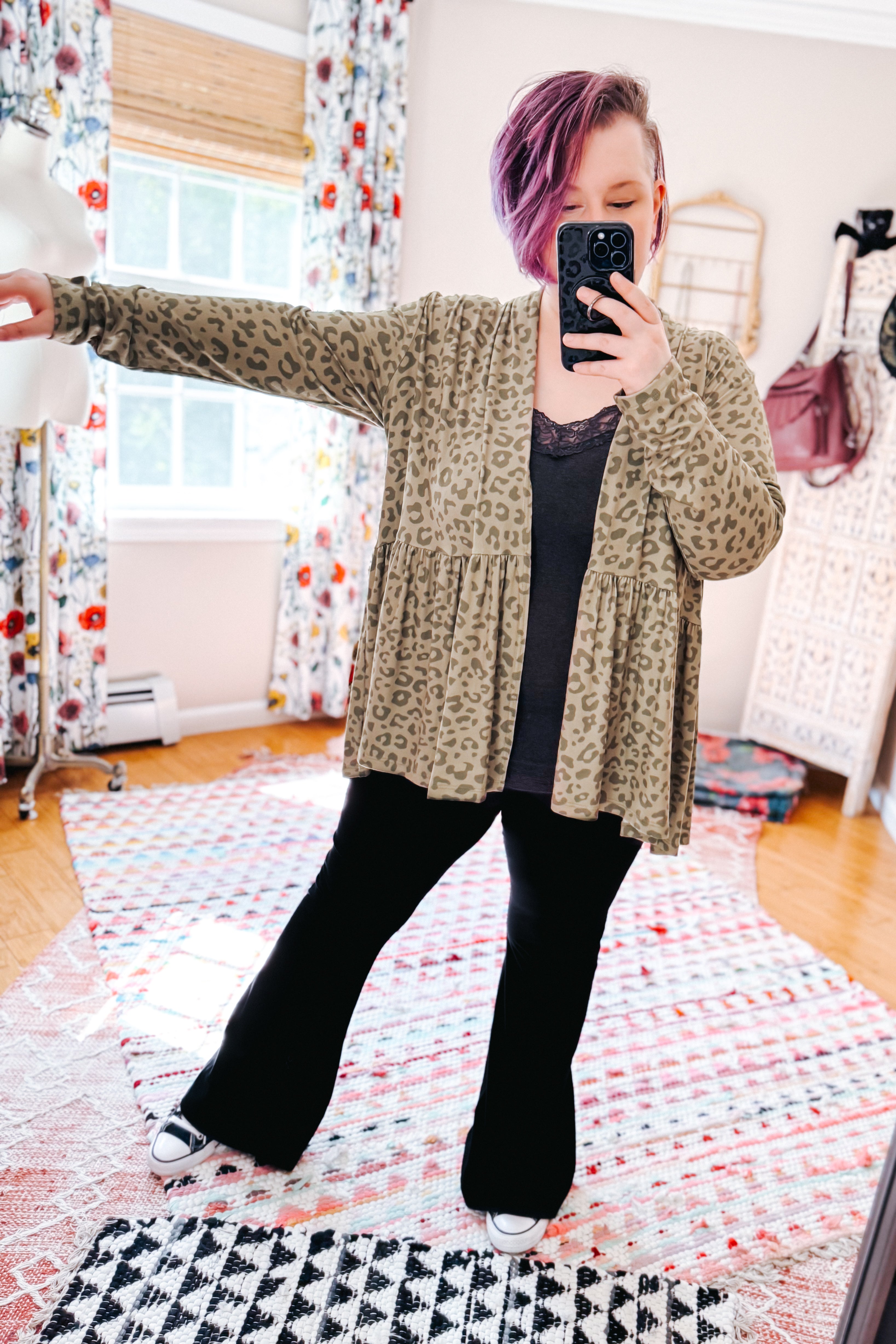 Woman with purple hair wearing open green leopard print peplum cardigan over black top and pants in bright eclectic room