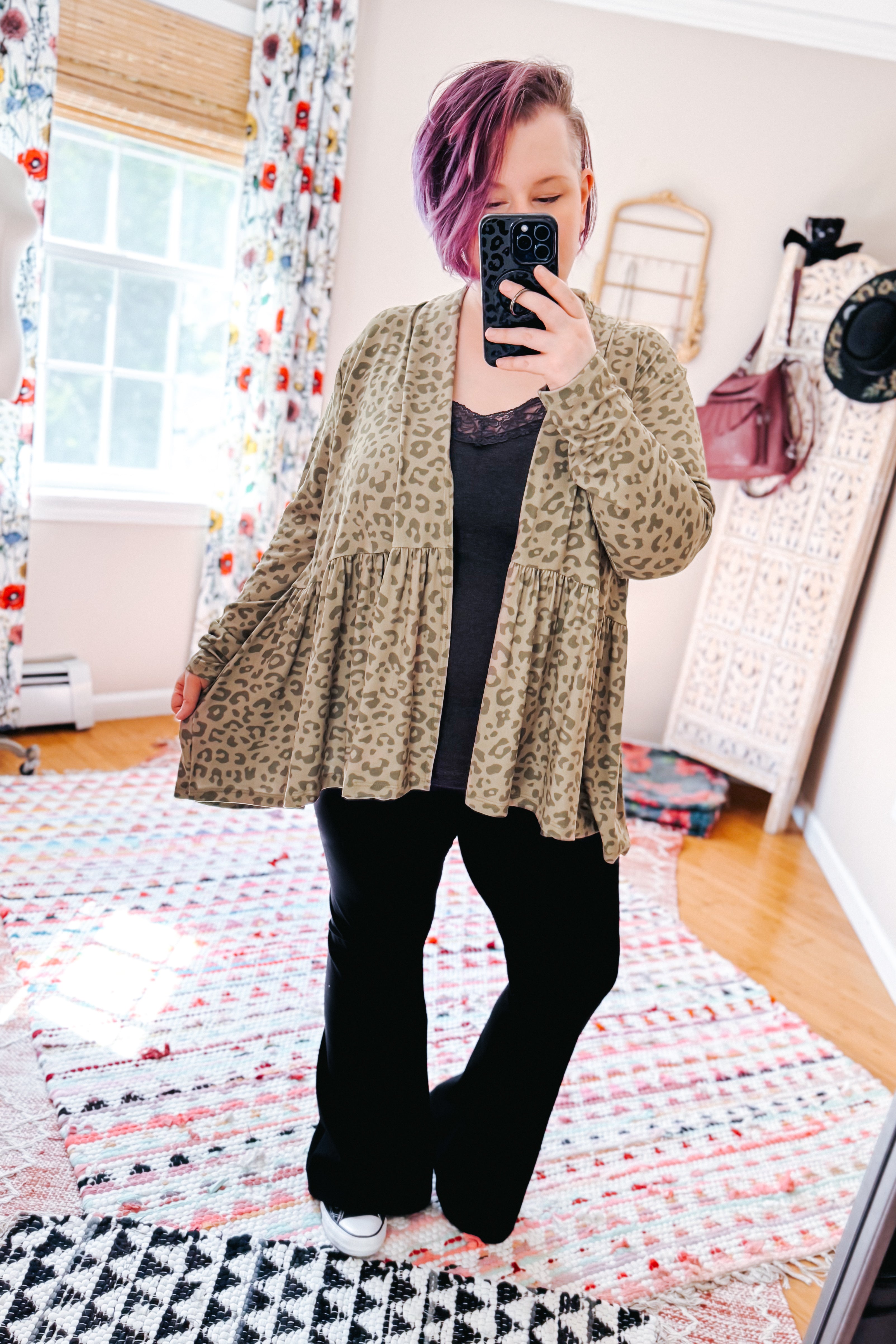 Woman with purple hair wearing green leopard print peplum cardigan over black top and pants in bright room with patterned rugs and decor