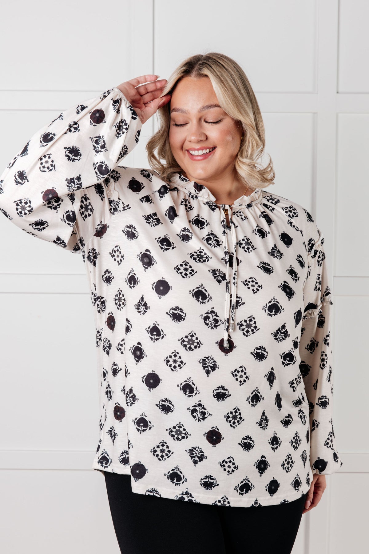 Smiling woman wearing a White floral tile blouse with a split neckline and ruffle details over black pants