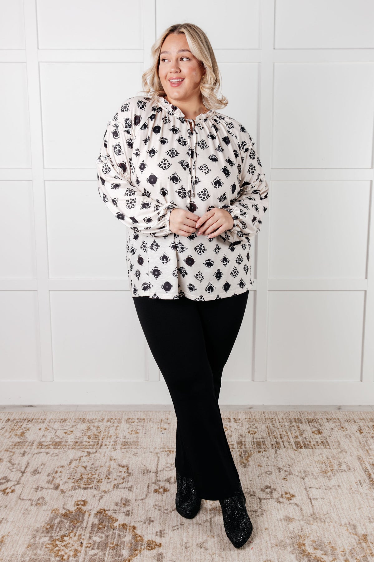 Smiling woman wearing a white floral tile blouse with ruffle details, black pants, and black shoes in a bright interior