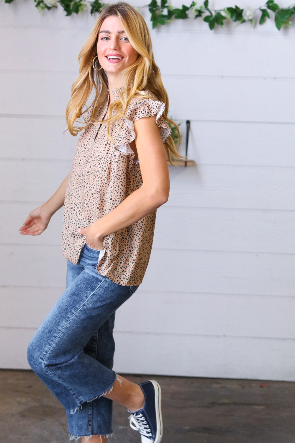 Woman wearing taupe animal print top with ruffle sleeves paired with cropped blue jeans and navy sneakers indoors