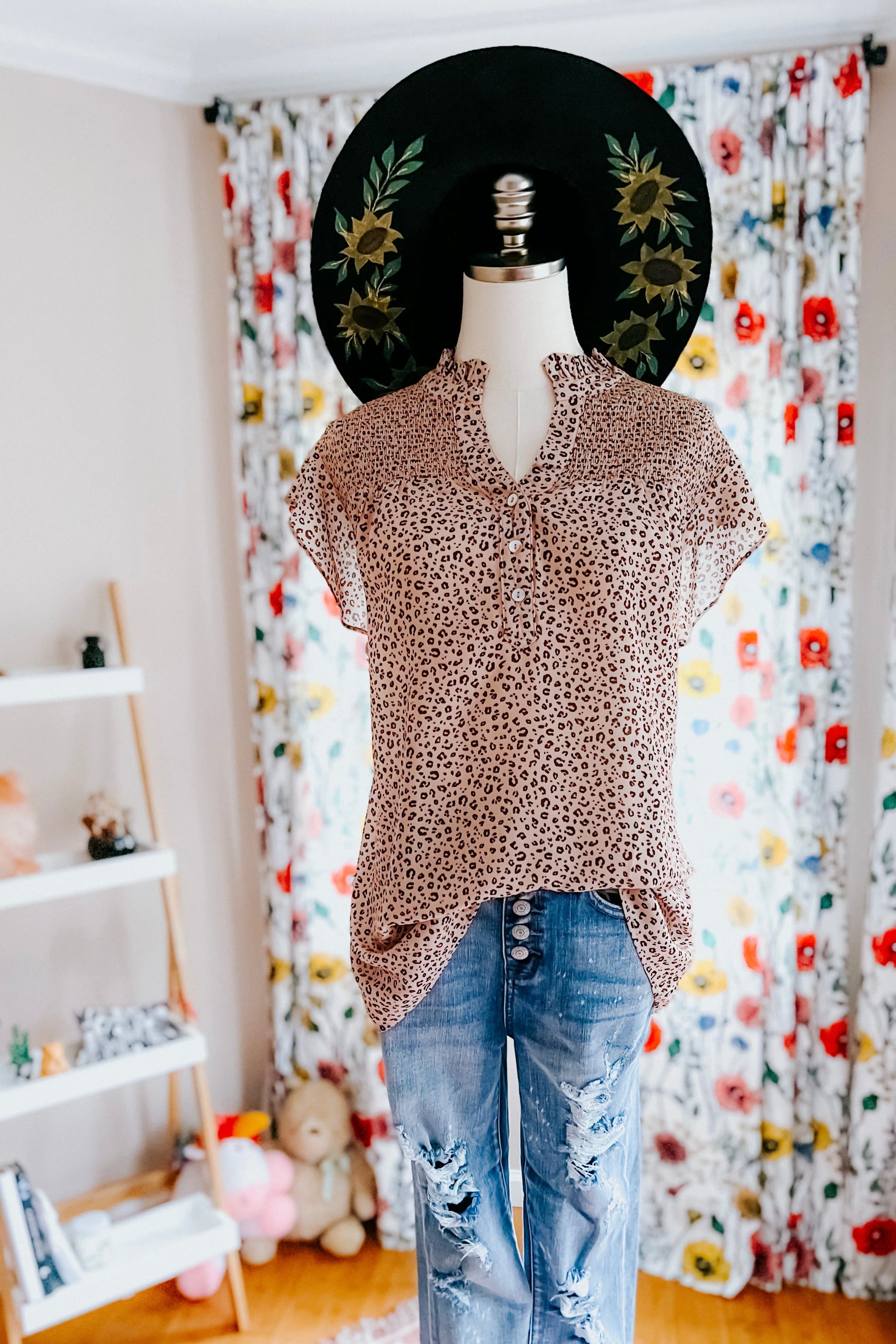 Taupe animal print blouse with ruffle collar styled with distressed high-rise jeans and black sunflower-decorated hat indoors