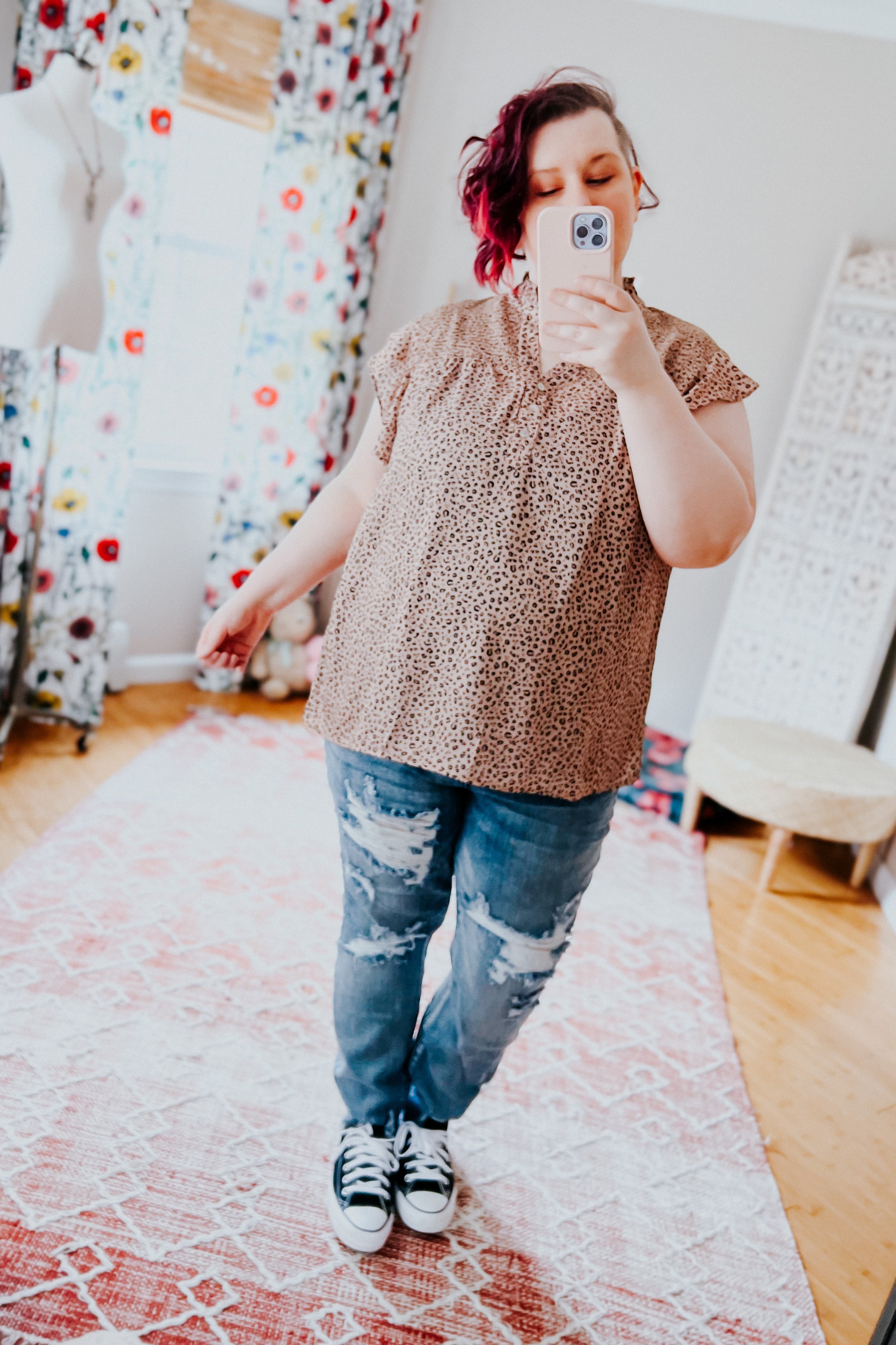 Woman wearing taupe patterned top with flutter sleeves paired with distressed jeans and black sneakers indoors