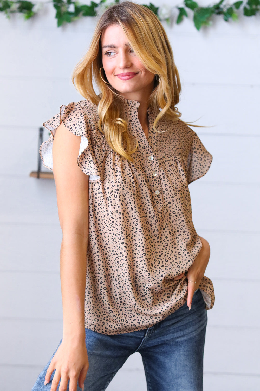 Woman wearing taupe animal print top with ruffled cap sleeves styled with blue jeans and gold hoop earrings indoors