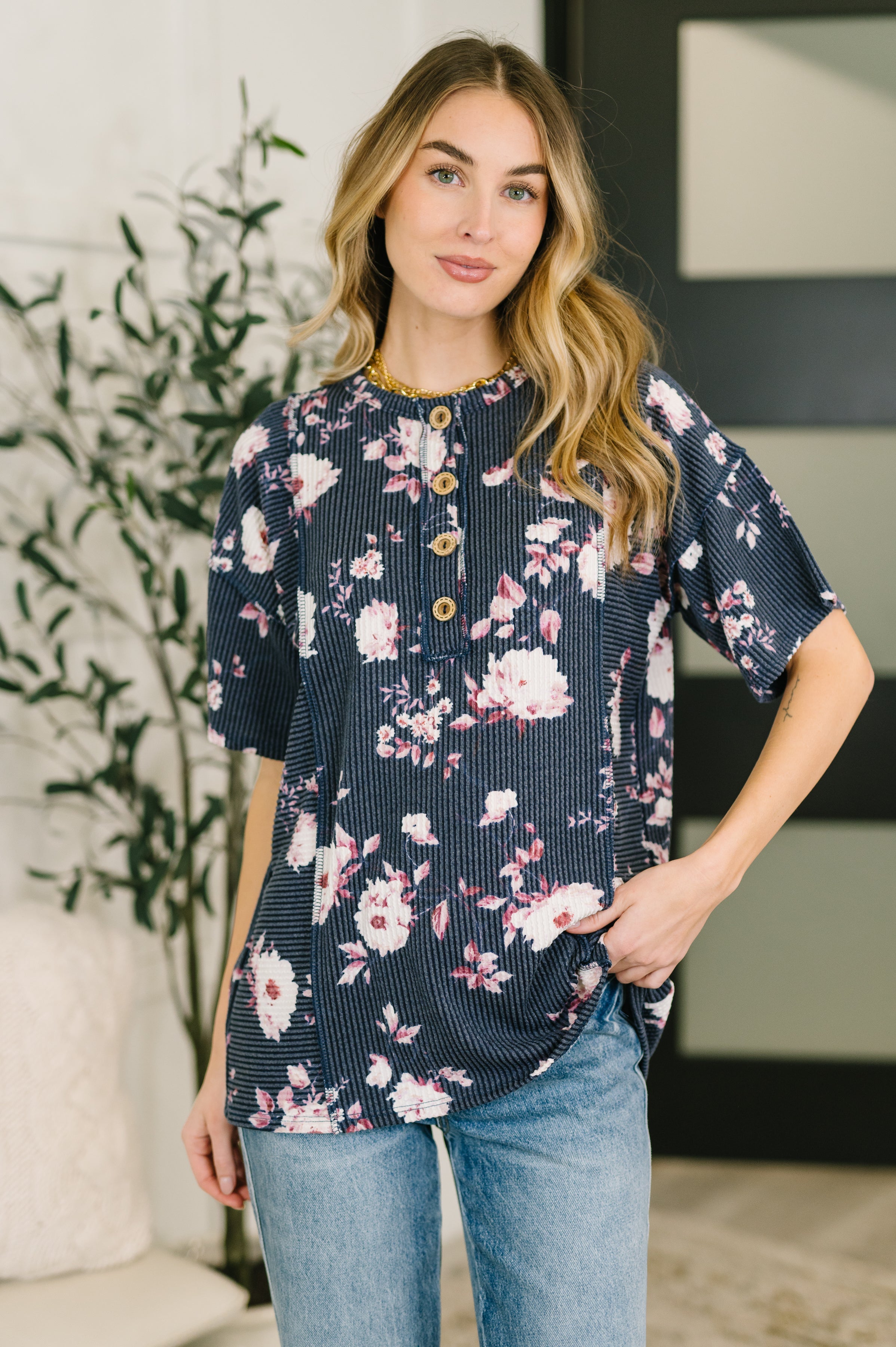 Woman wearing a navy floral top with short sleeves and wooden buttons styled with light wash jeans indoors