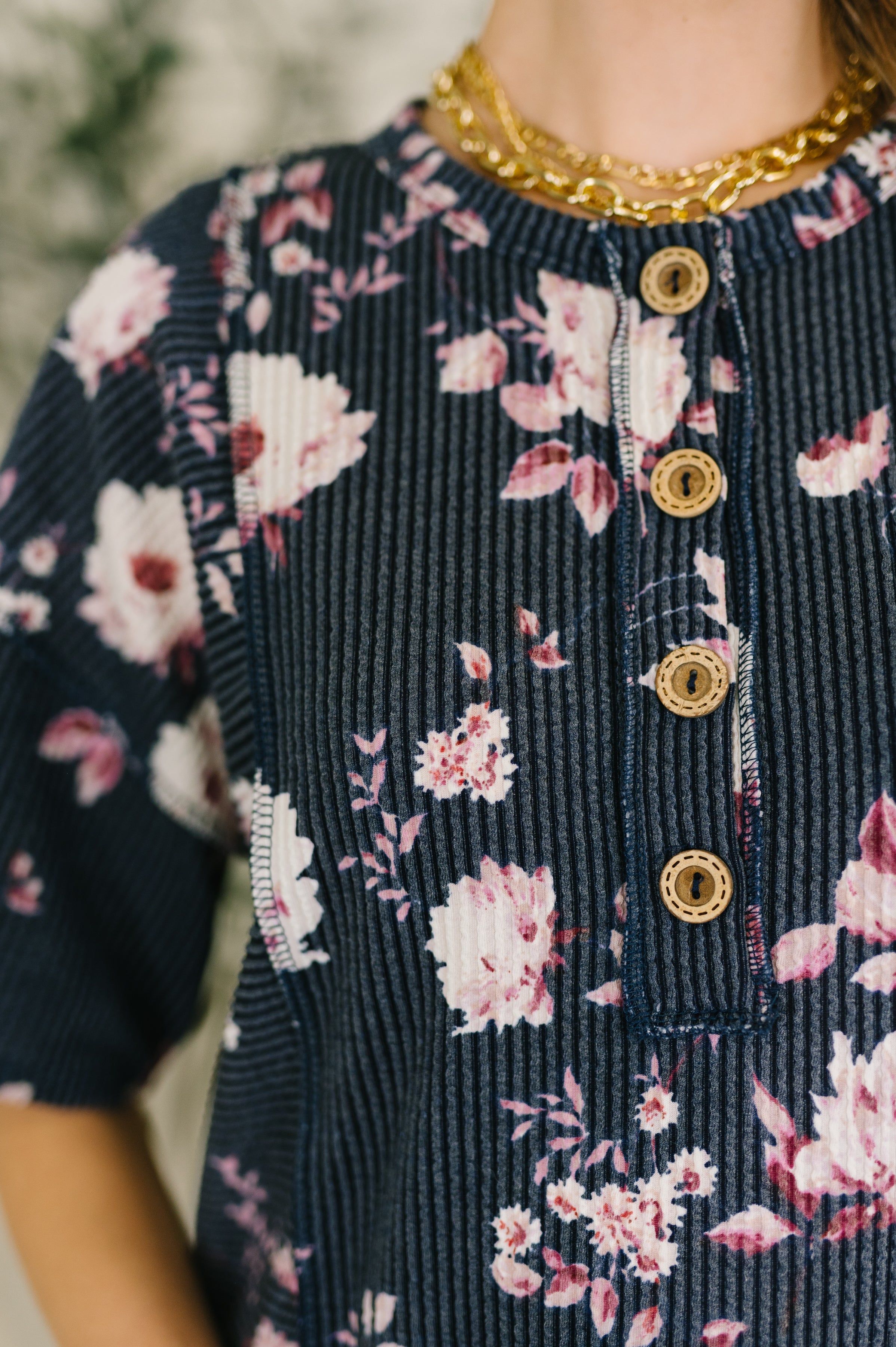 Close-up of navy textured top with pink floral print and wooden buttons worn with layered gold chain necklaces