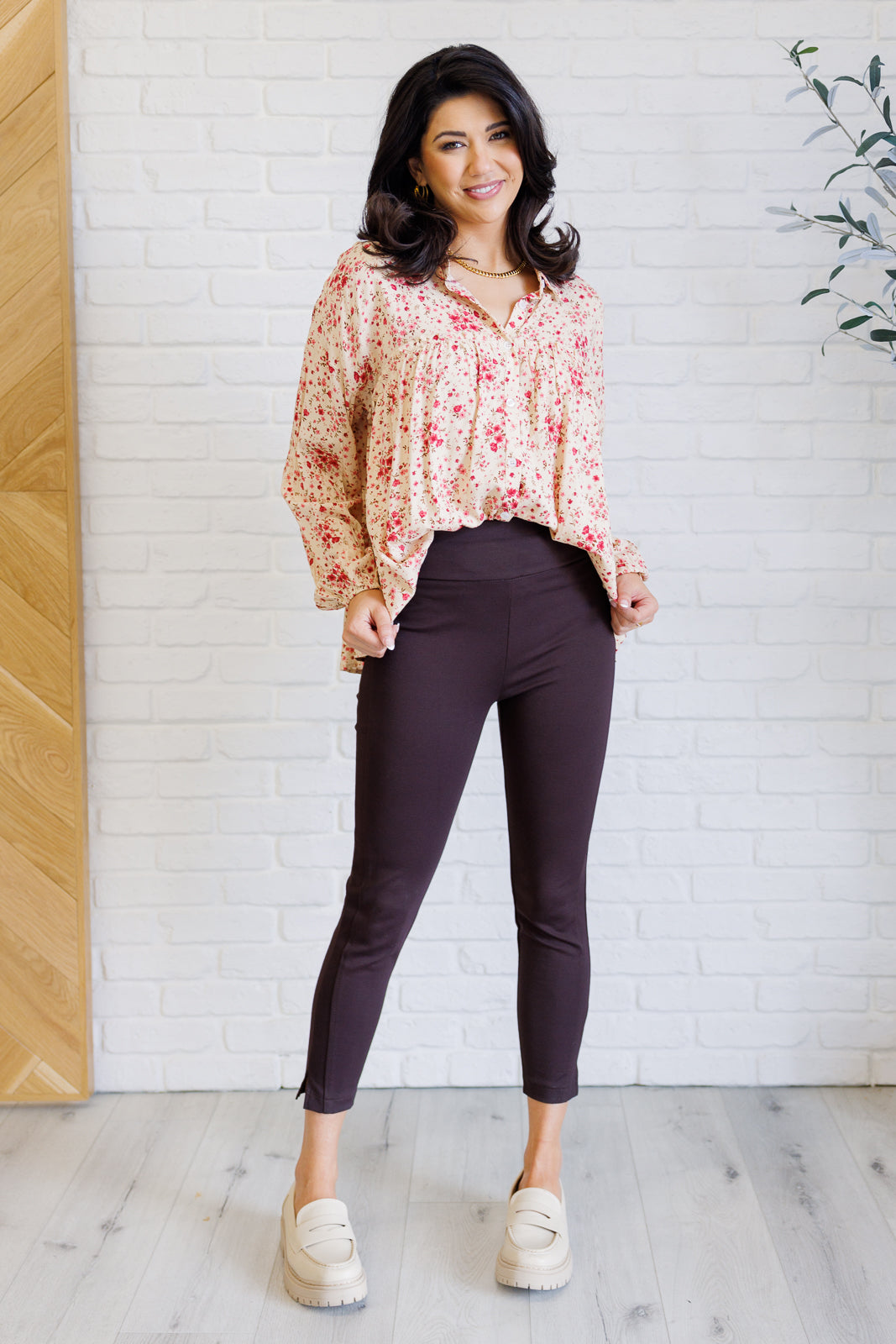 Smiling woman wearing a pink floral button down shirt with balloon sleeves over dark fitted pants and platform shoes indoors