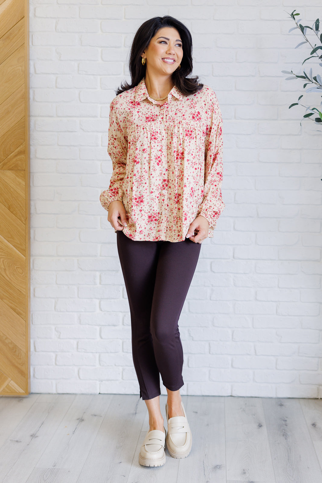 Smiling woman wearing a pink floral collared button down shirt with long sleeves and dark leggings and loafers indoors
