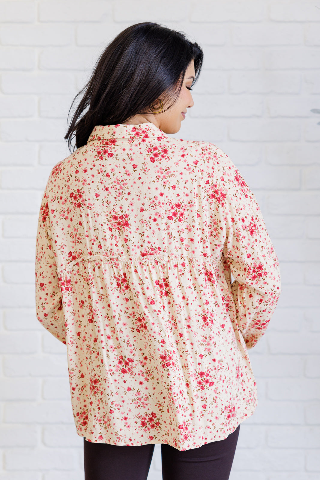 Woman viewed from behind wearing a pink floral collared shirt with relaxed balloon sleeves and dark pants against a white brick wall