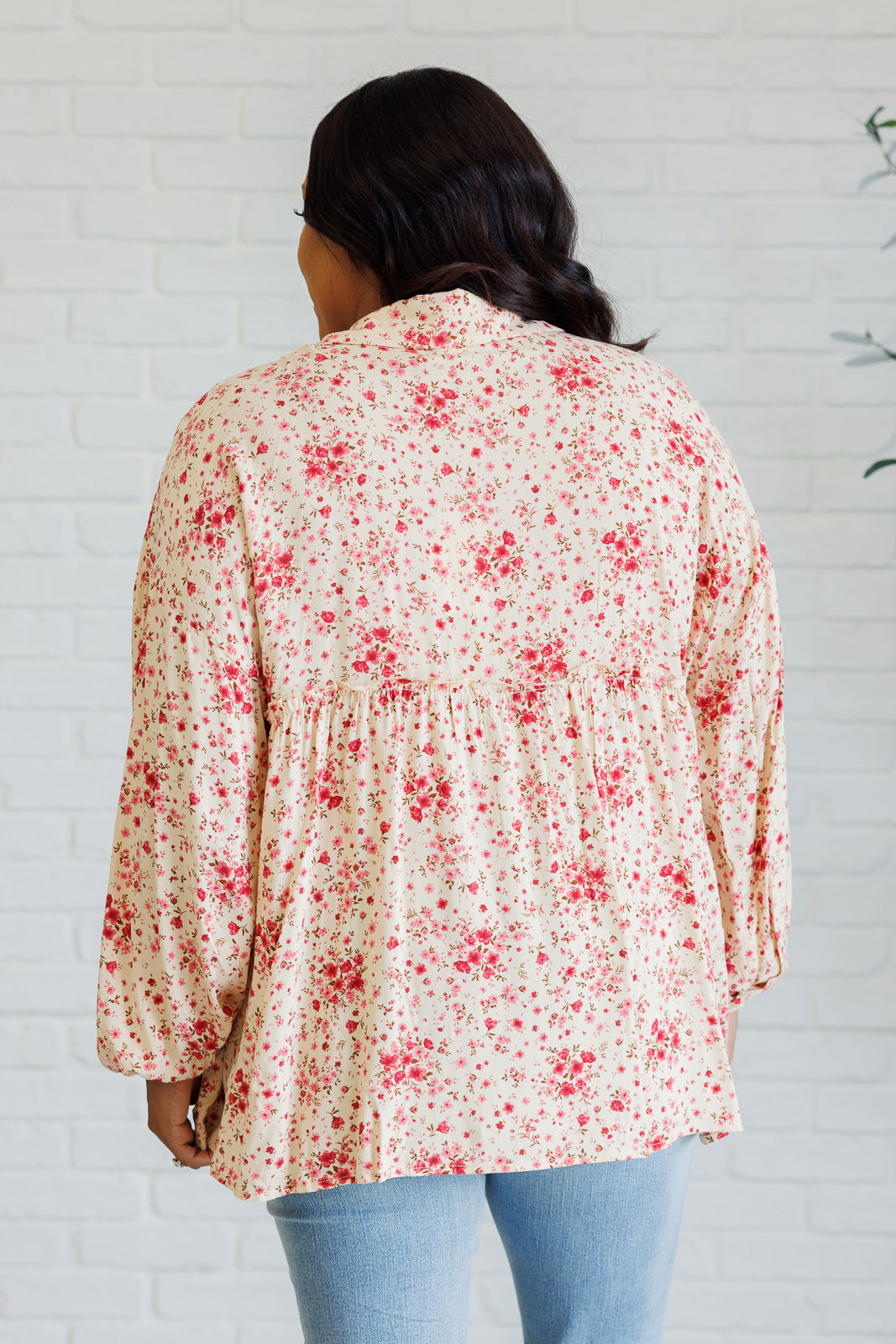 Woman seen from behind wearing a pink floral button down shirt with balloon sleeves and a gathered waist over light blue jeans indoors