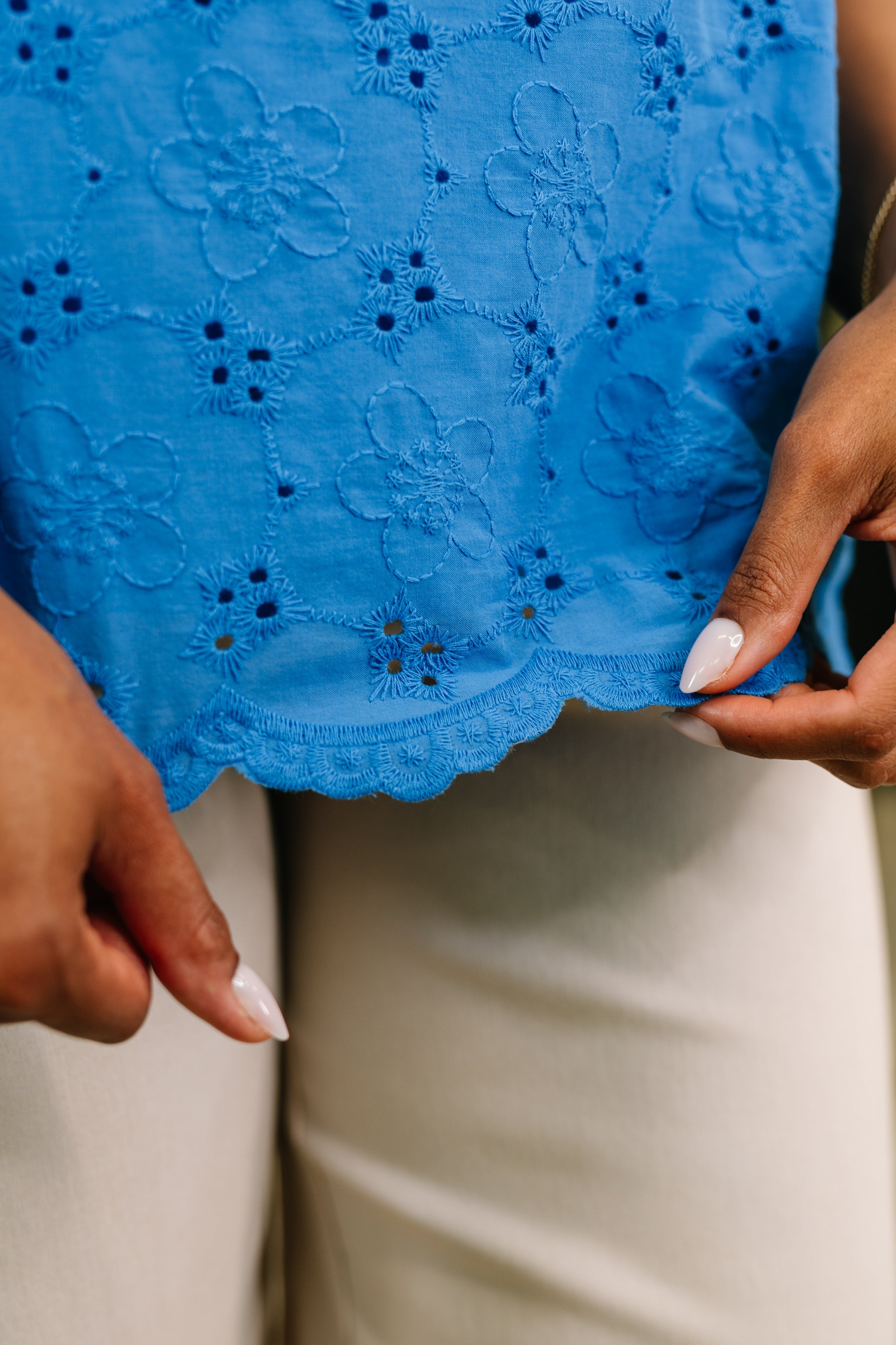 Close-up of blue eyelet fabric with floral embroidery and scalloped edge held by hands with neutral nails