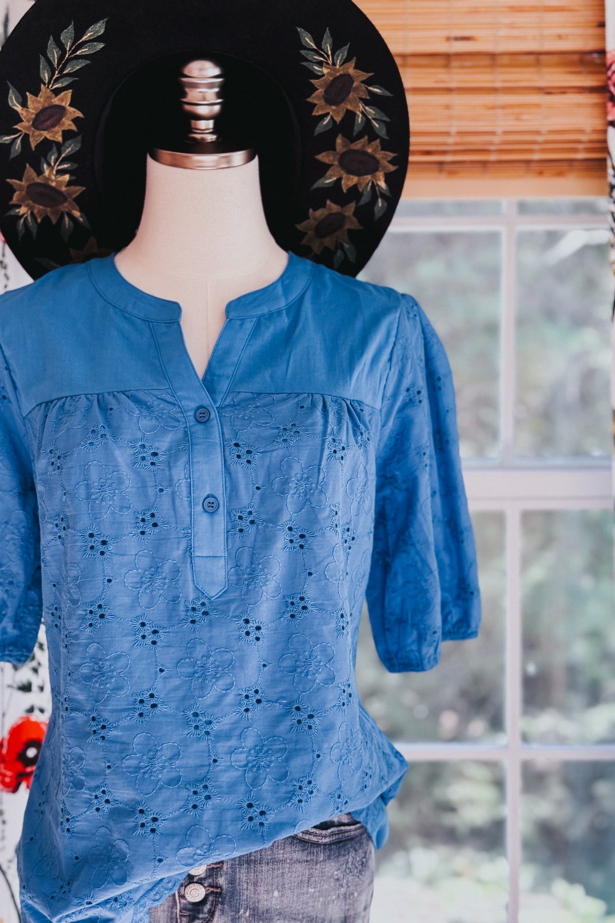Blue eyelet blouse on a mannequin with embroidered floral cutout details a split neckline and three quarter sleeves indoors