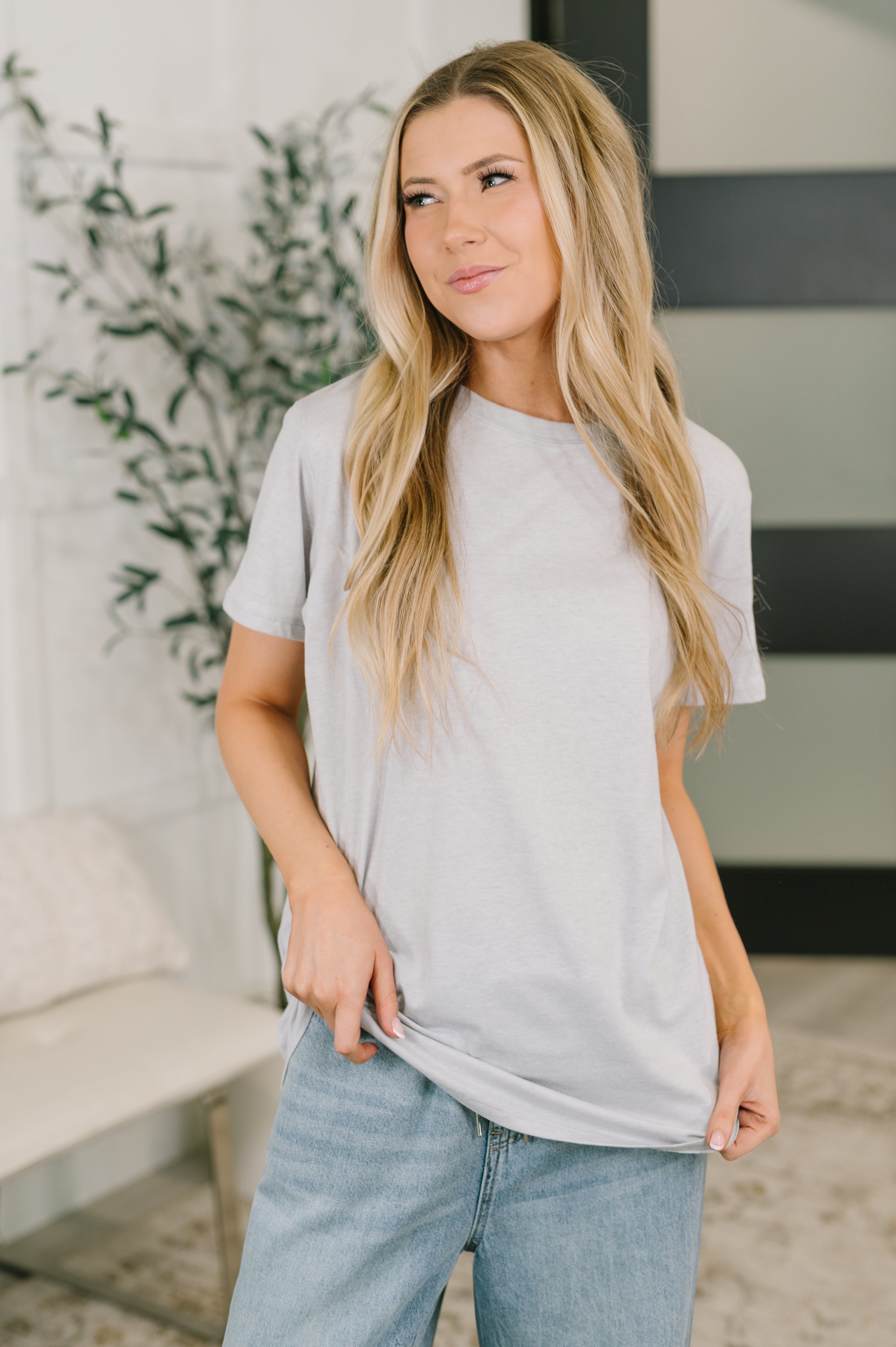 Woman wearing heather grey relaxed fit tee with short sleeves paired with light blue jeans in cozy room