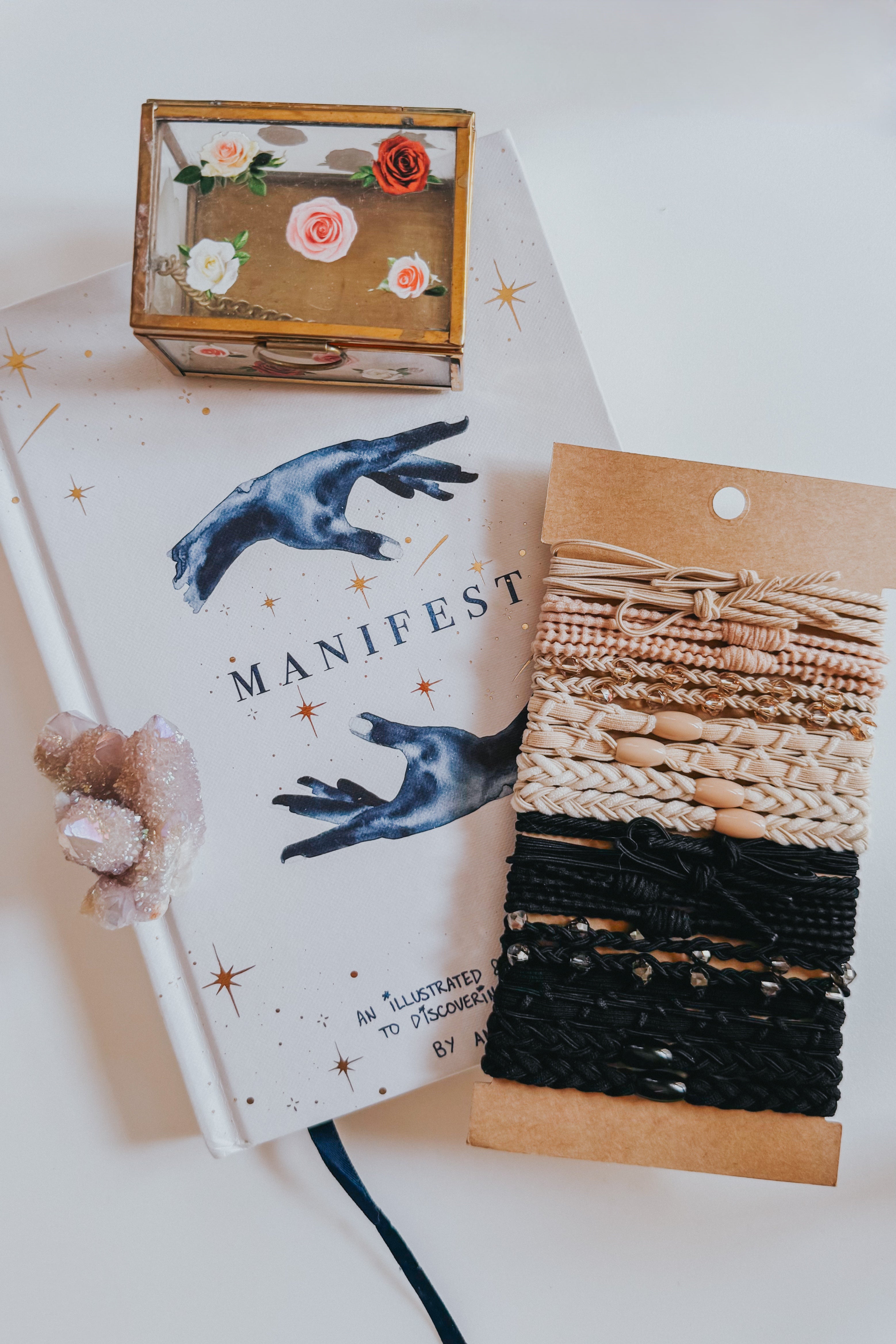 Set of cream and black woven stackable hair tie bracelets with bead and braided details on brown card backing beside a decorative box and book