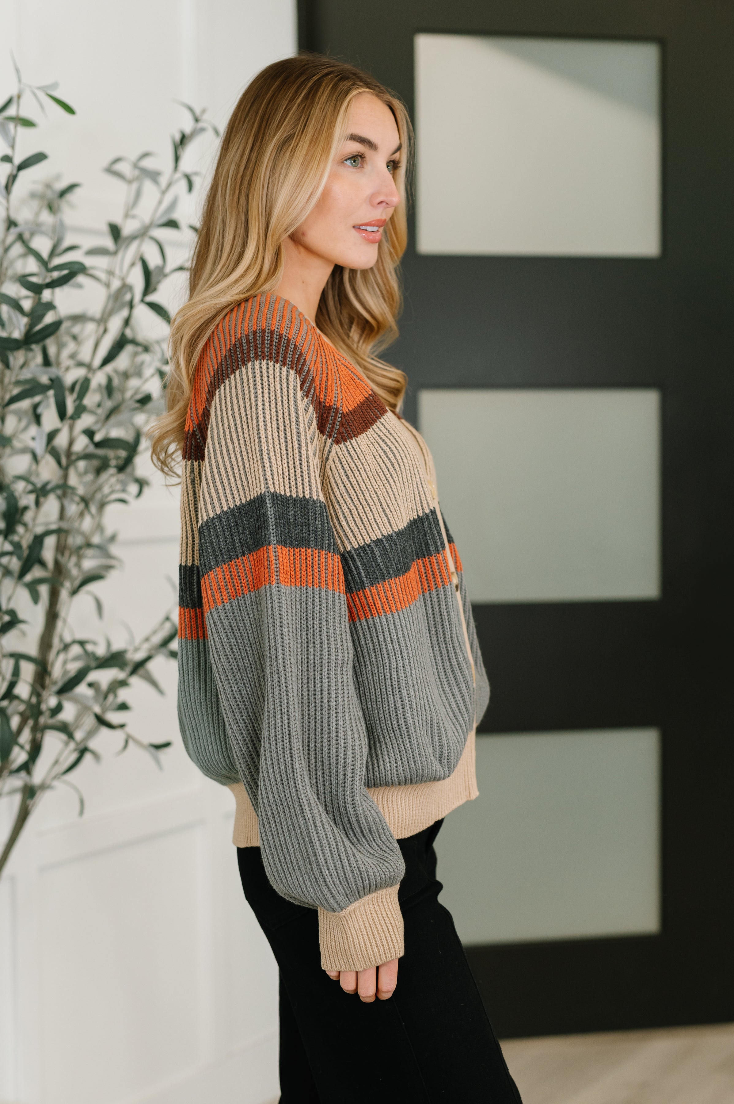 Side view of woman wearing a relaxed tan cardigan with orange, gray, and rust stripes paired with black pants indoors