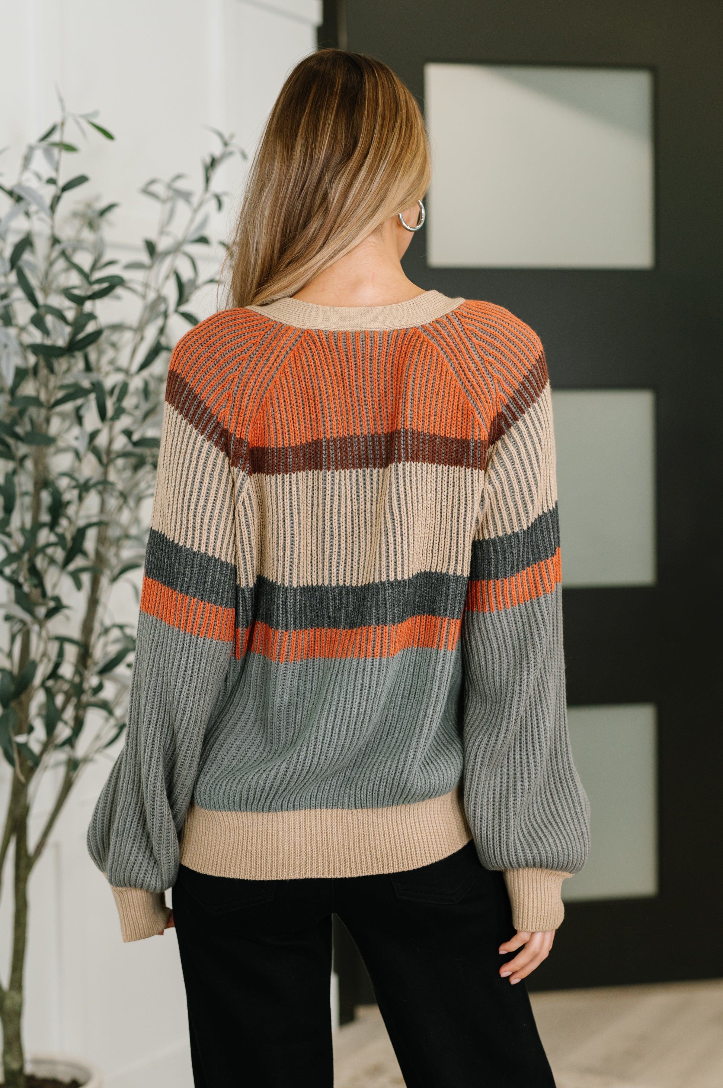 Back view of woman wearing a relaxed tan knit cardigan with bold orange, brown, and gray stripes paired with black pants indoors