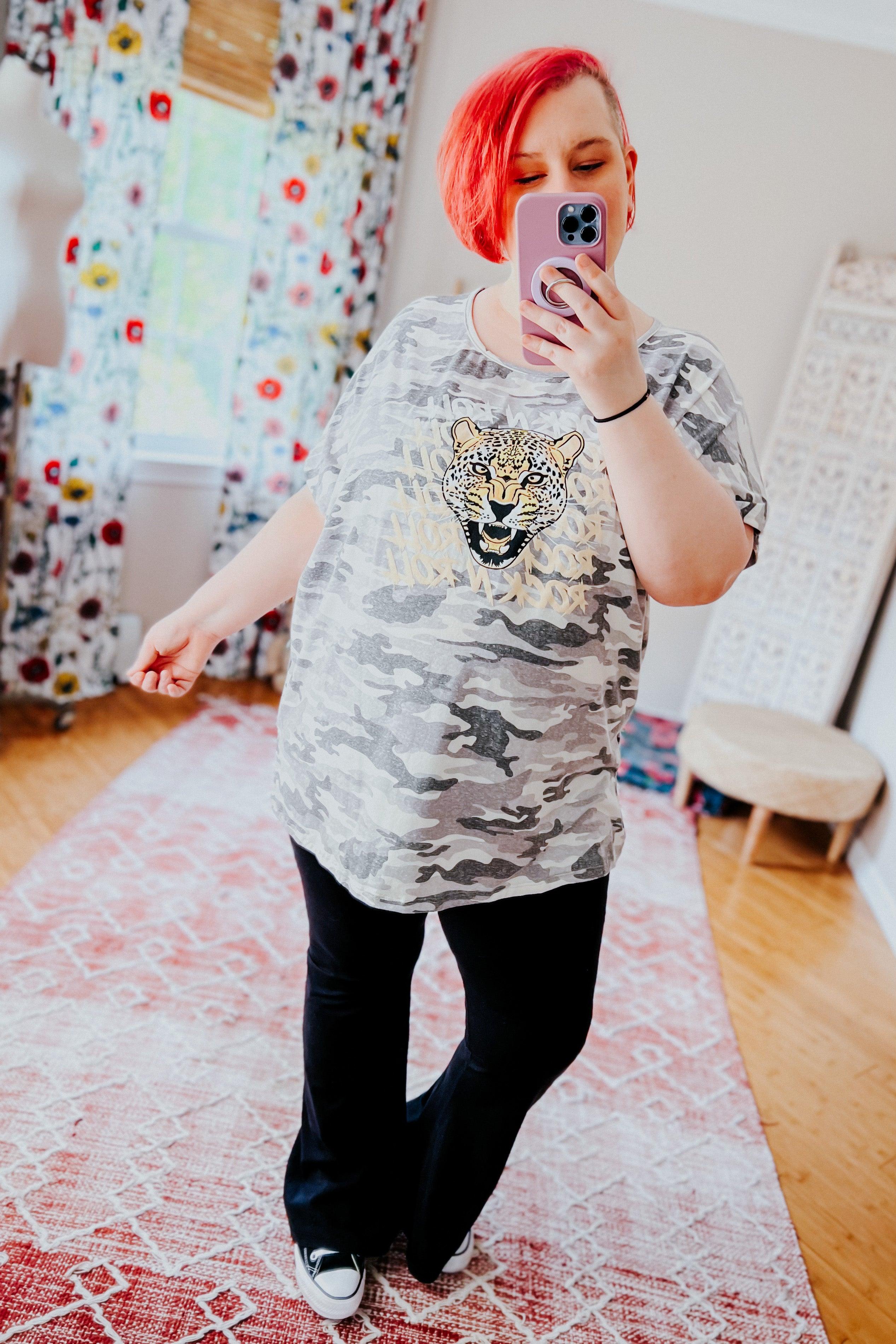 Woman with bright red hair wearing grey camo tee with leopard graphic and black pants taking mirror selfie in bright room