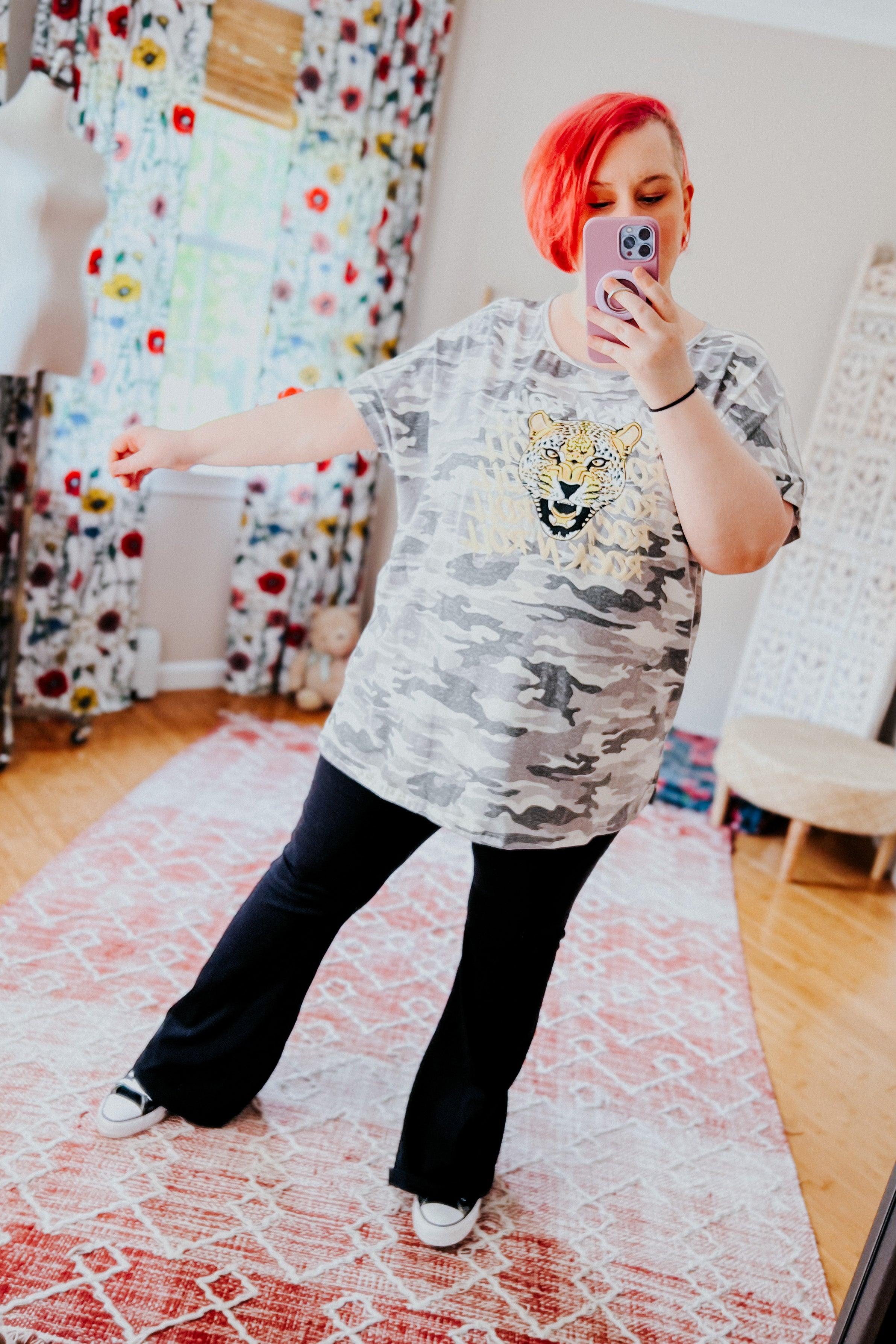 Plus size woman with bright red hair wearing grey camo tee with bold leopard graphic and black flare pants taking selfie in bright room