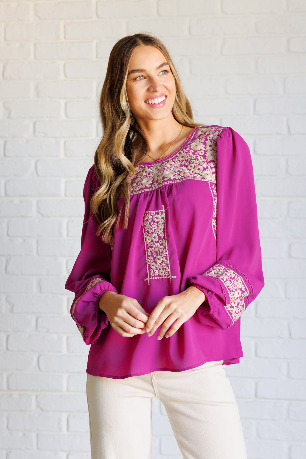 Woman wearing a magenta long-sleeve blouse with intricate white embroidery and beige jeans standing against a white brick wall.
