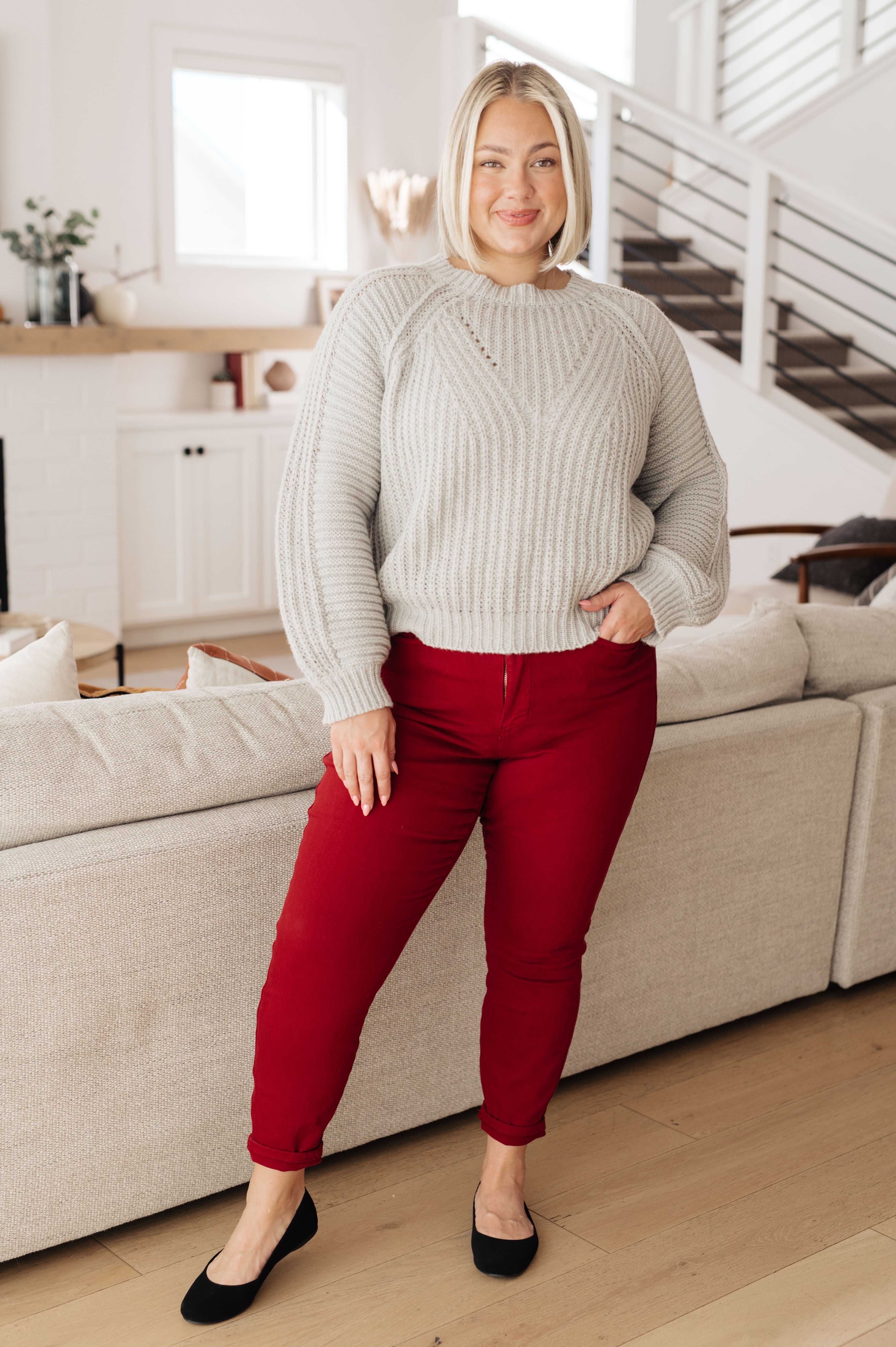 Plus size woman smiling wearing grey balloon sleeve sweater and red pants standing in bright living room