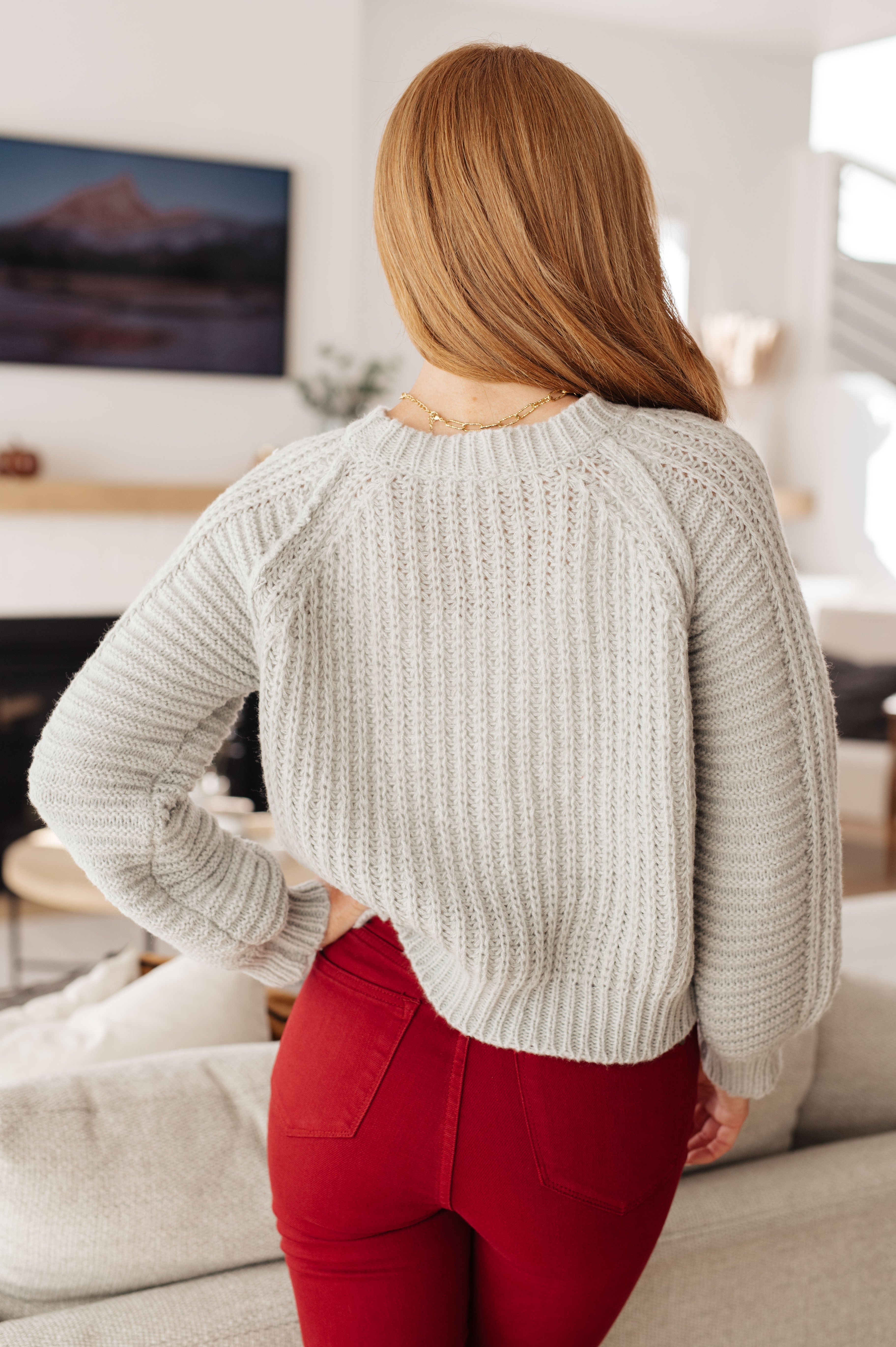 Woman with long light brown hair wearing grey knitted balloon sleeve sweater and red pants standing indoors