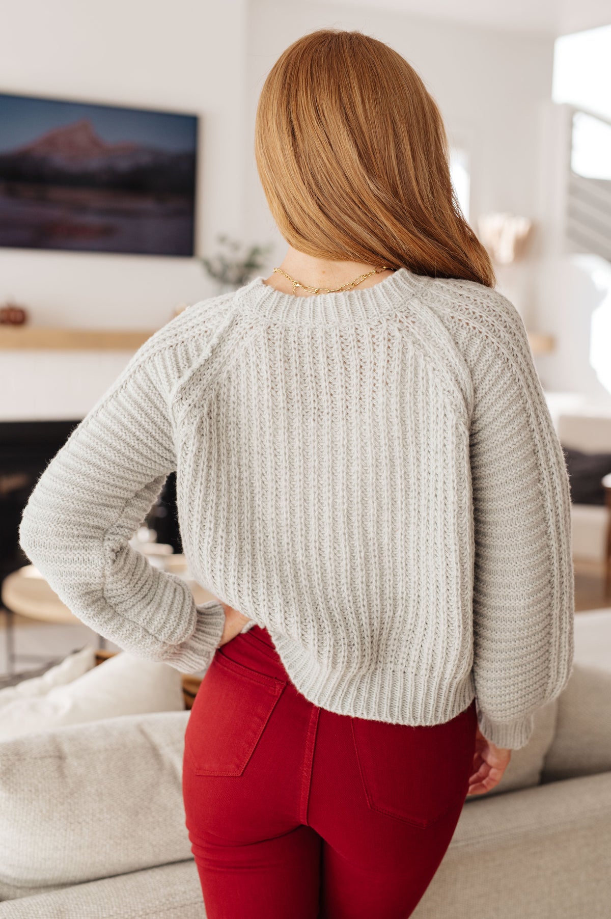 Back view of a woman wearing a grey ribbed sweater with balloon sleeves and red pants in a bright living room