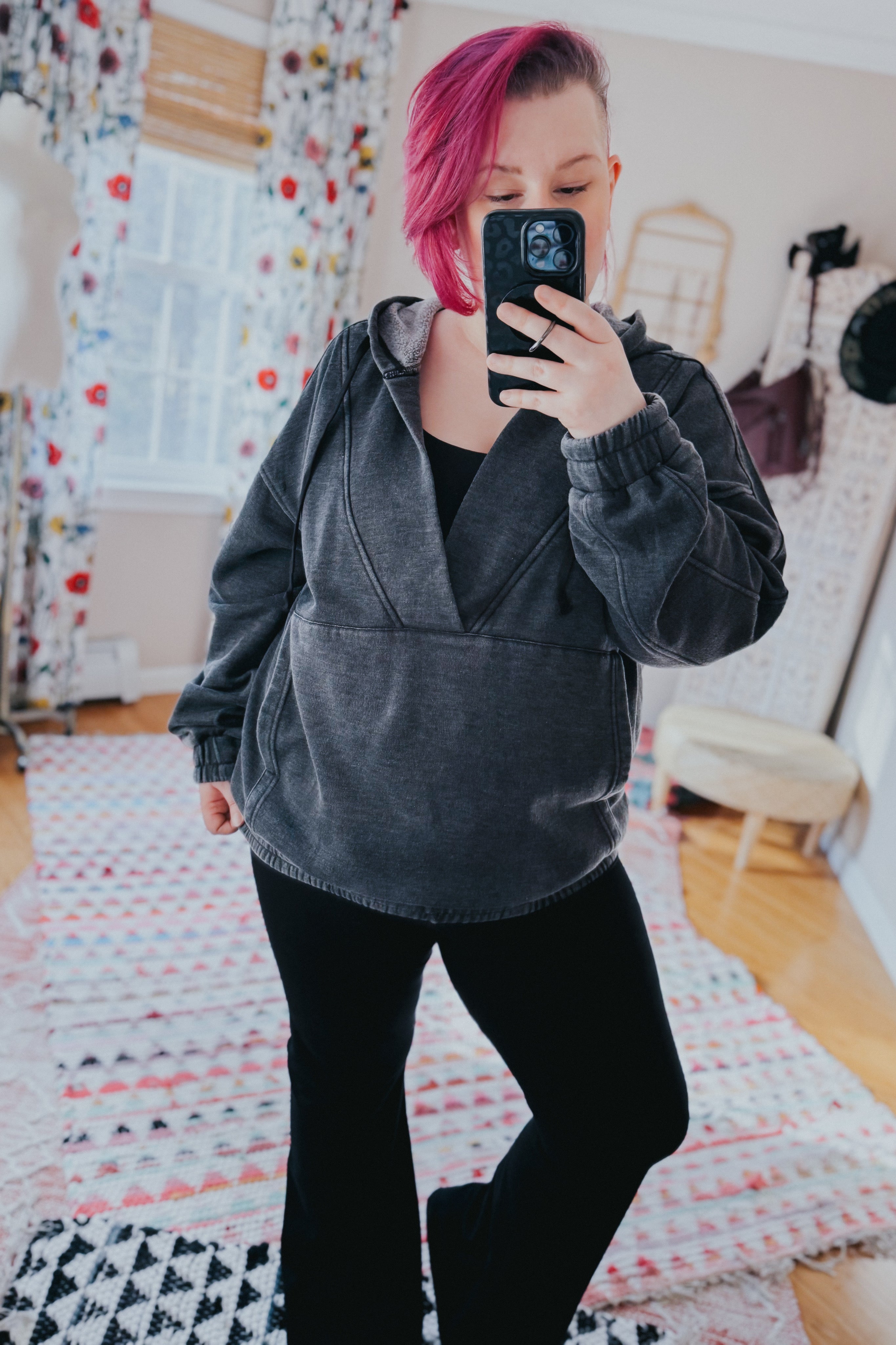 Woman with short magenta hair wearing charcoal acid wash hoodie and black pants taking mirror selfie indoors