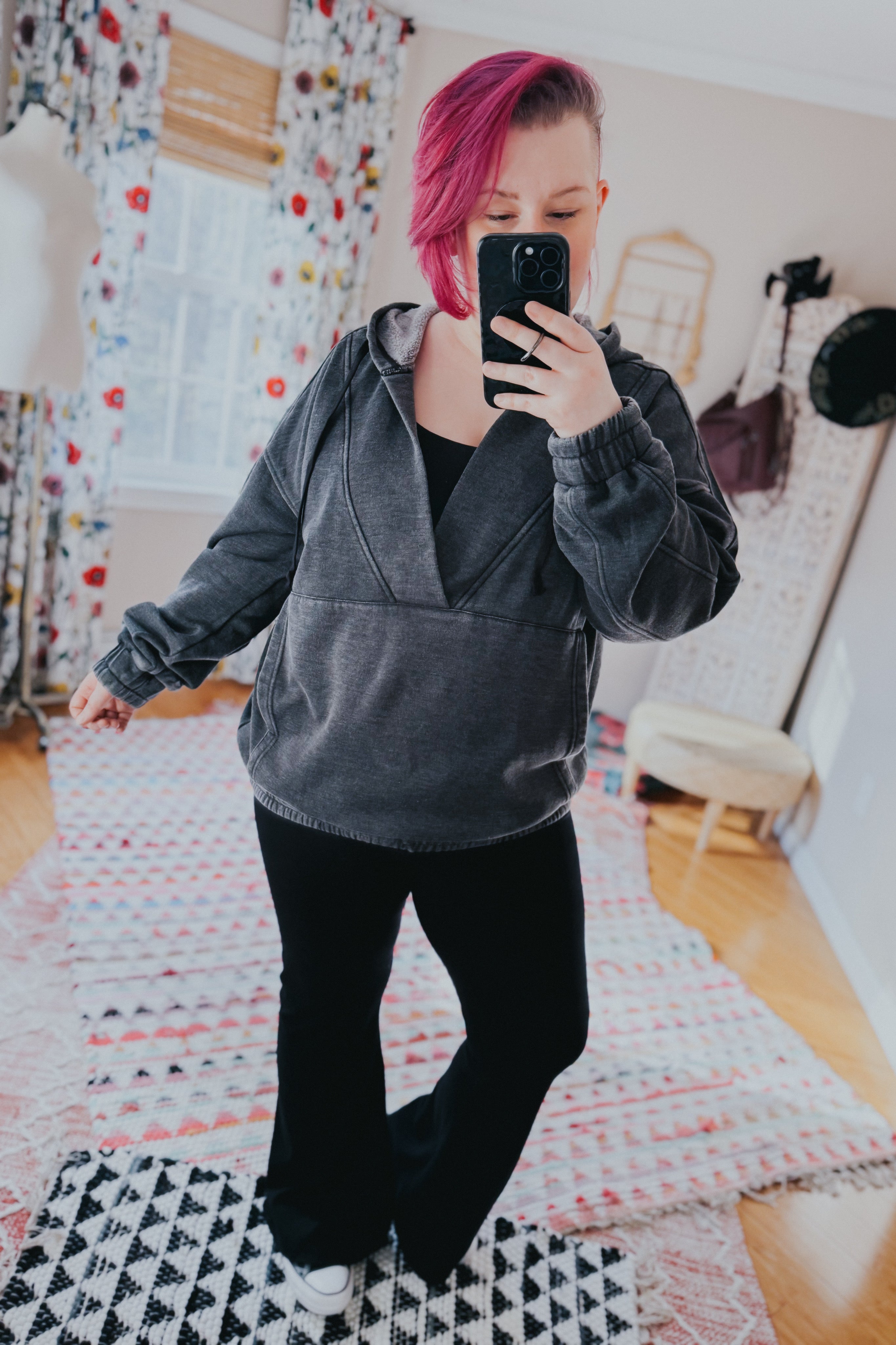 Woman with pink hair wearing charcoal acid wash hoodie styled with black pants taking mirror selfie indoors