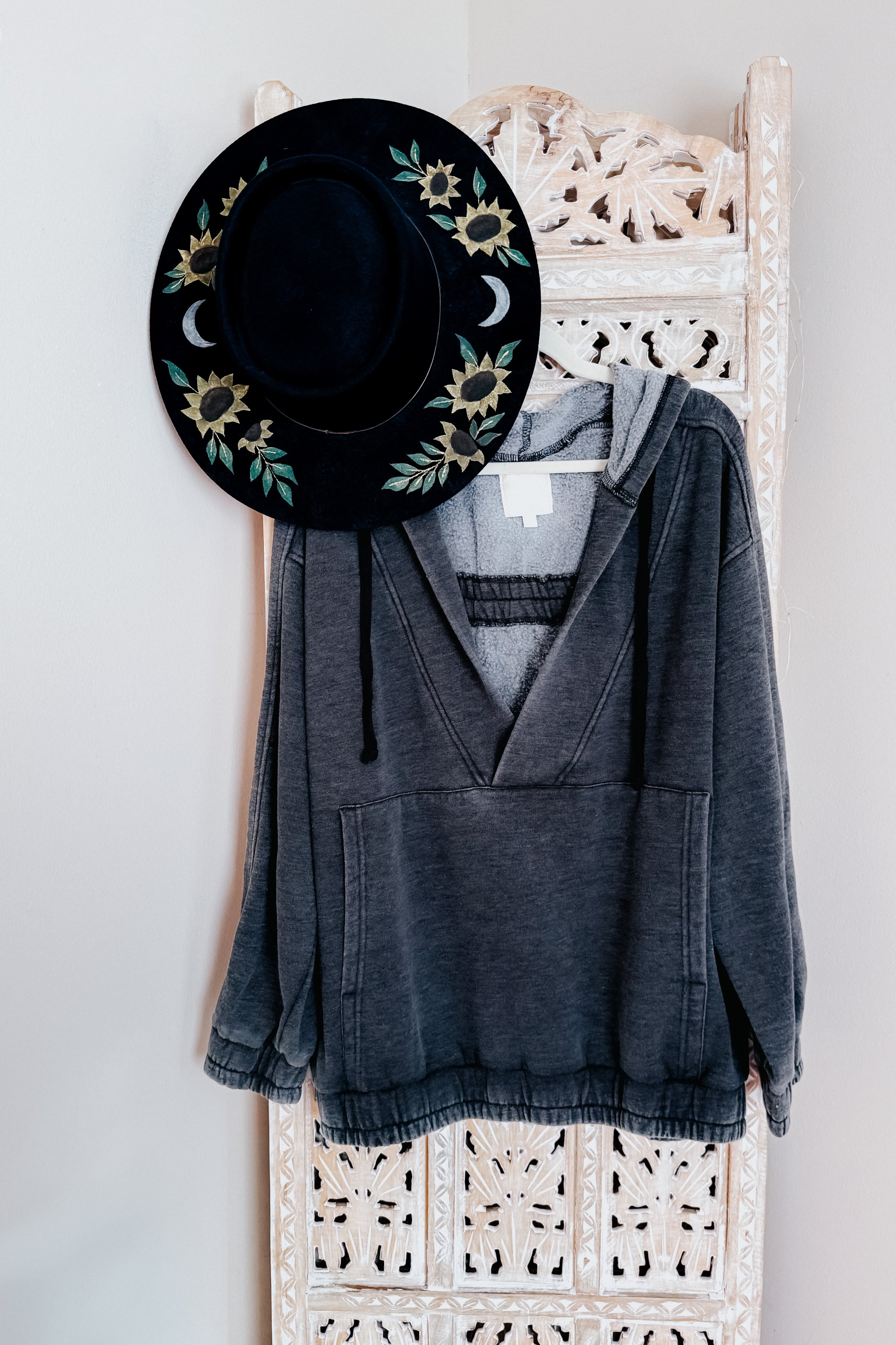 Charcoal acid wash hoodie hanging on ornate white wooden panel with black wide-brim hat featuring sunflower and moon designs